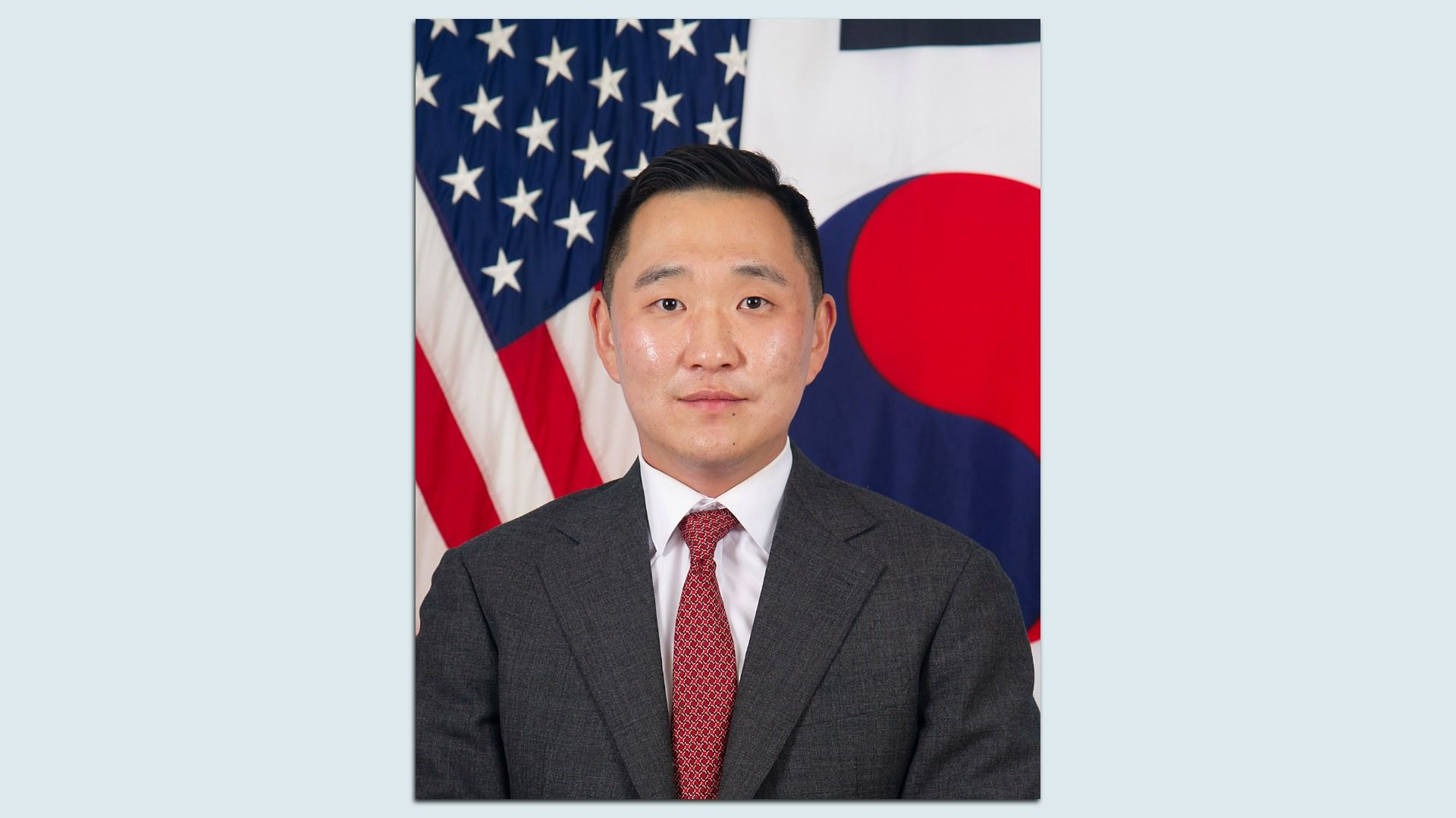 Portrait of an Asian man in a dark gray suit and red tie, standing in front of the United States and South Korea flags.