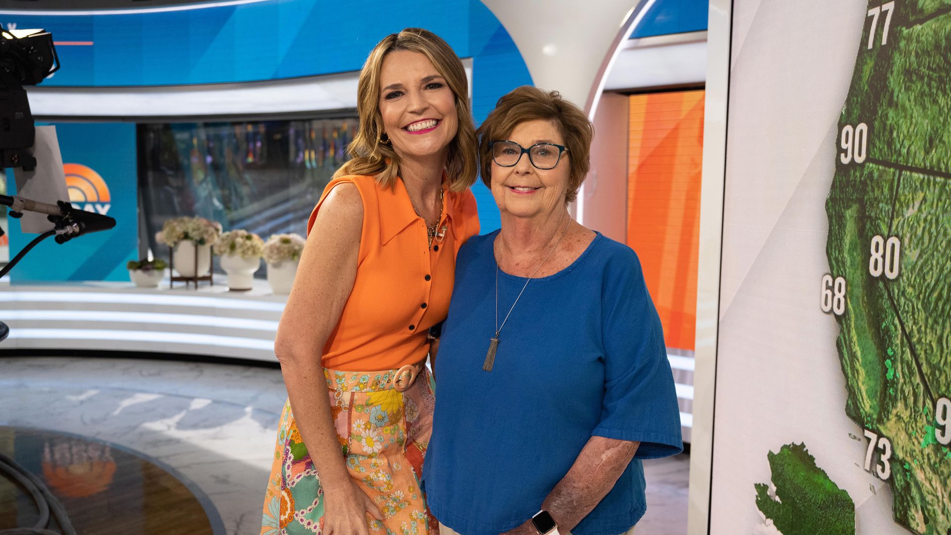 Two women smiling in a TV studio; one in an orange sleeveless top with patterned skirt, the other in a blue shirt and beige pants, standing beside a weather map showing temperatures.