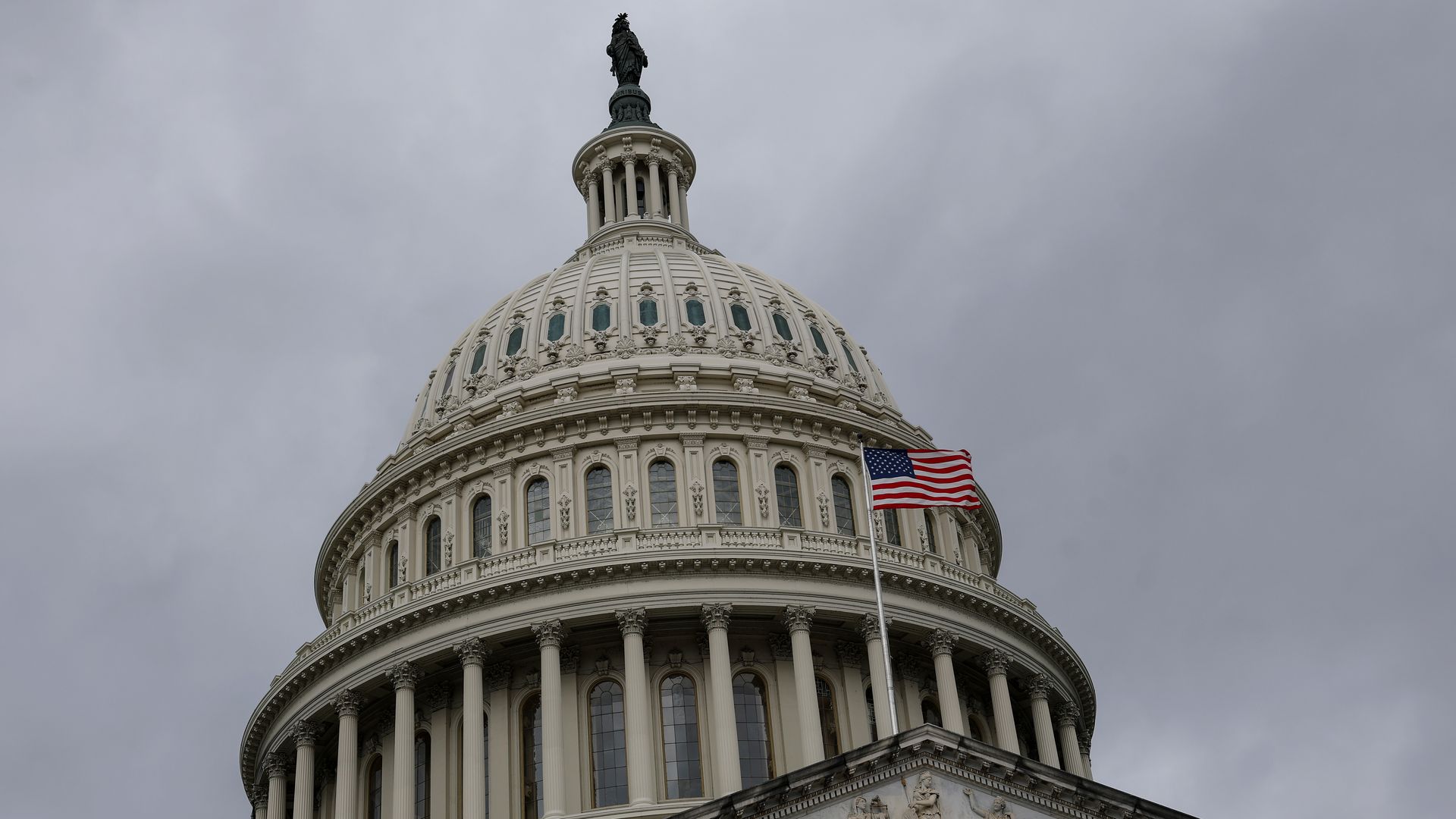 White neoclassical Capitol dome with statue atop, tall flagpole bearing the American flag in front, ornate pediment and columns beneath, against a gray overcast sky.