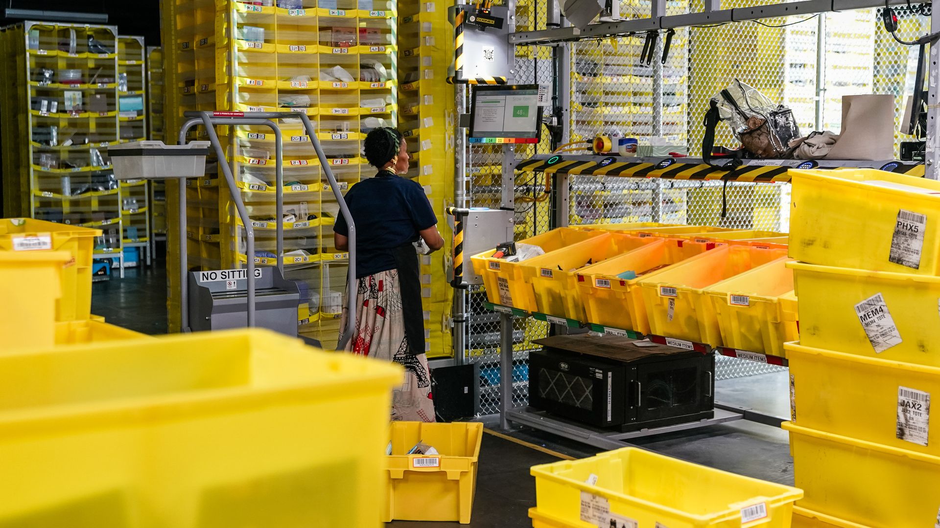 Warehouse with numerous yellow bins and crates, a person in a patterned skirt and dark shirt working near bins, shelves filled with small items.