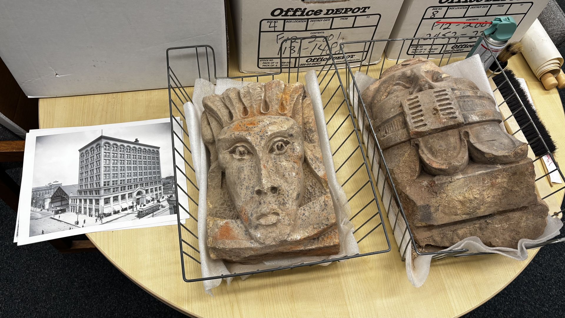 Two weathered gray stone sculptures in metal racks on a wooden table: a carved head with a headdress on the left and a layered helmet on the right; a black-and-white photo sits nearby.