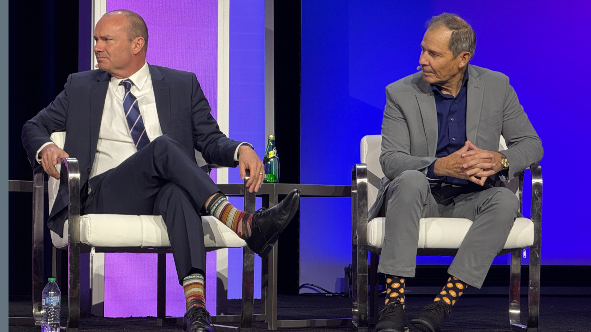 Two men in suits on stage, seated, at a conference, with an LED screen behind them in blue and purple.