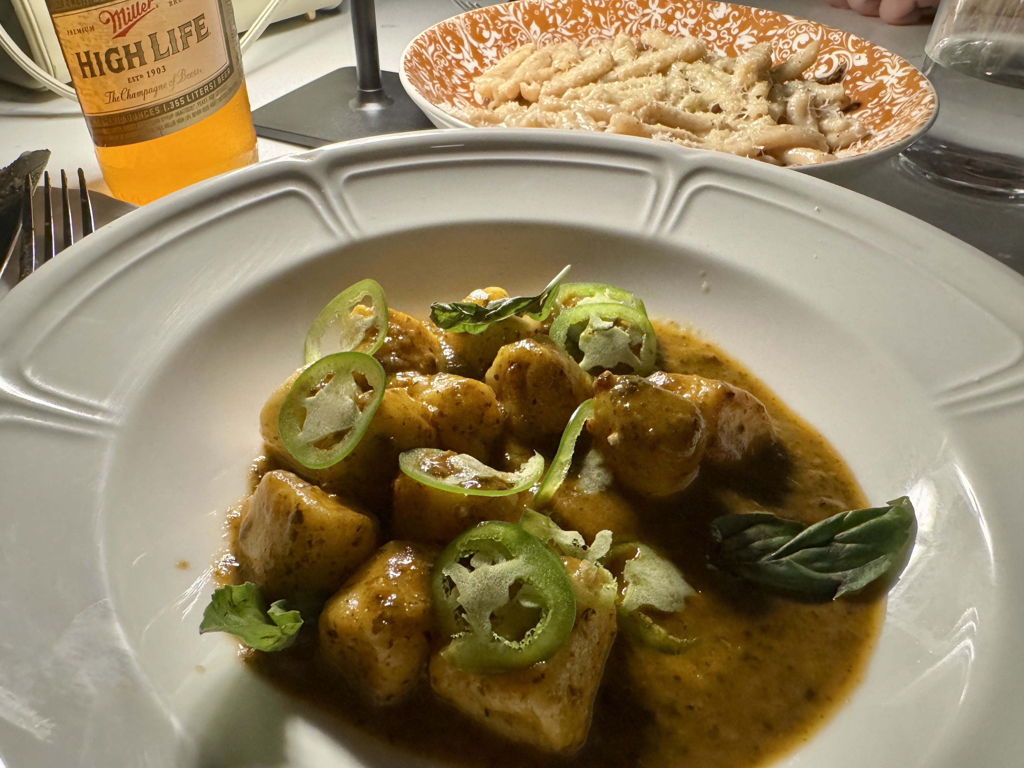 Plated gnocchi in brown sauce garnished with green chili slices and basil leaves on a white plate; background shows a bowl of pasta with grated cheese and a bottle of Miller High Life beer.