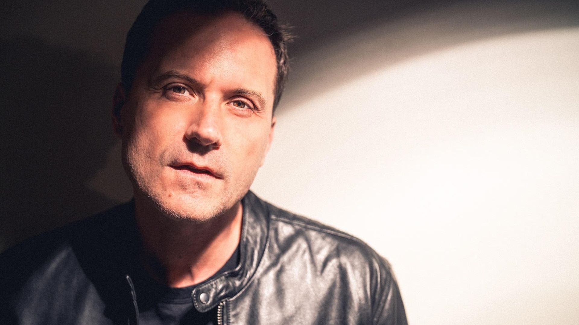 Zayd Ayers Dohrn with short dark hair in a black leather jacket, facing the camera with a subtle smile. The left side is dim, while a bright circular light appears on the right.