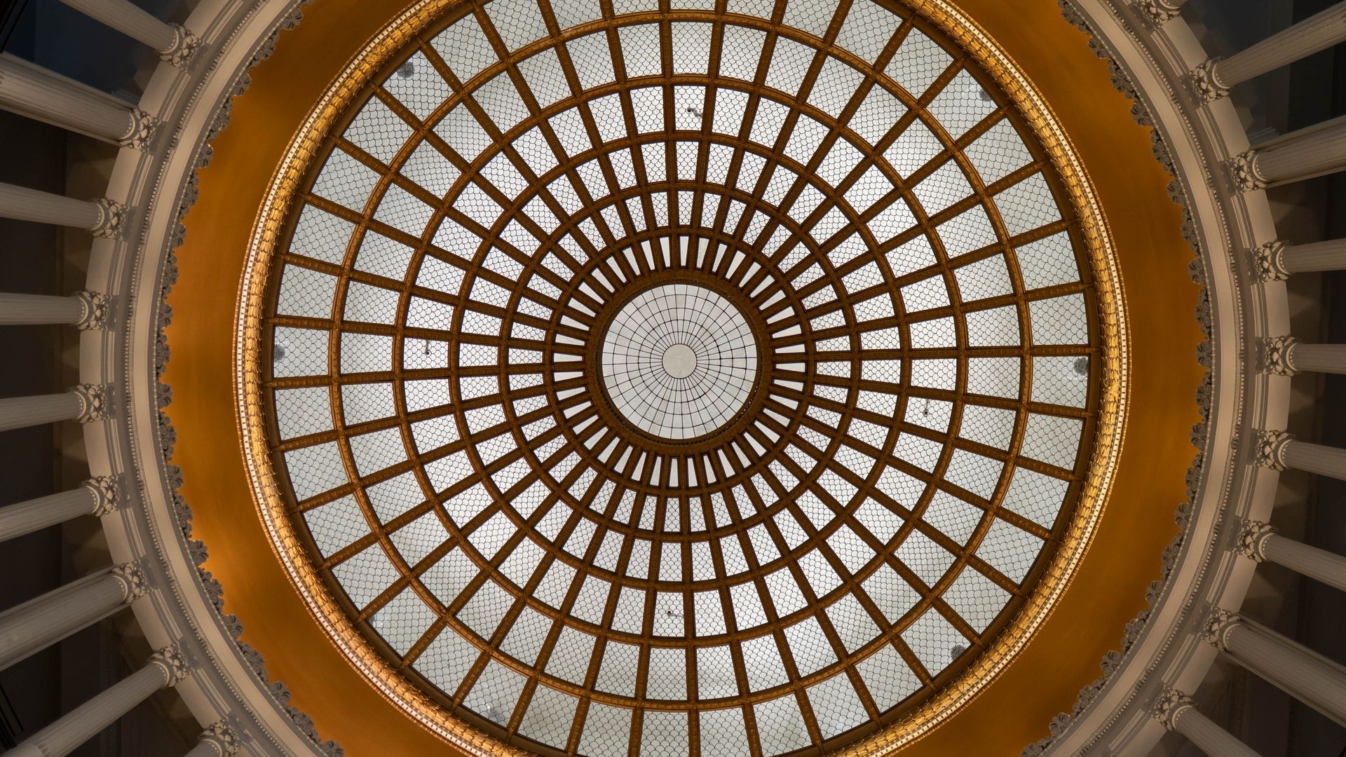 Looking up at a grand circular glass rotunda with a gold lattice rim and radiating spokes, surrounded by a white ornate colonnade along the dome’s edge.