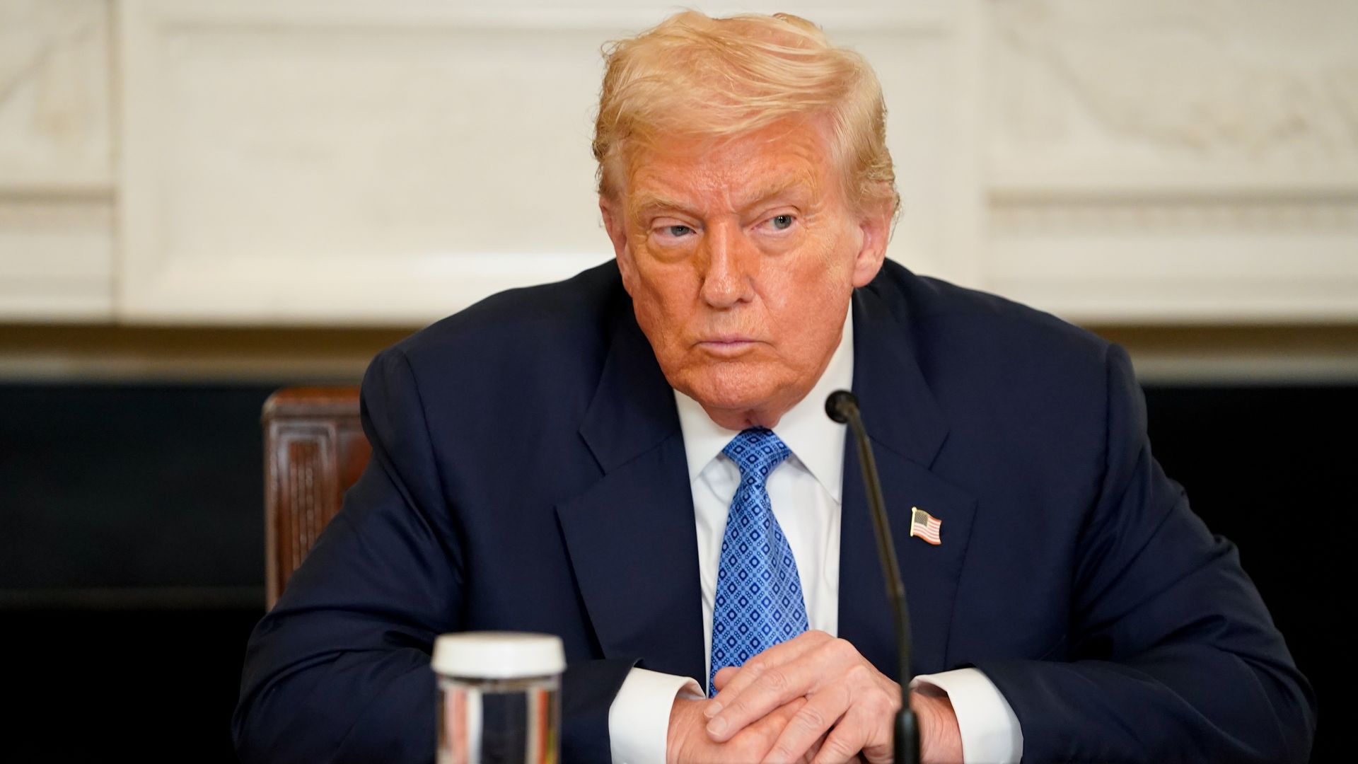 A serious man with light orange hair in a navy blue suit and blue tie sits at a table with hands clasped, an American flag pin on his lapel, and a microphone and water glass in front.