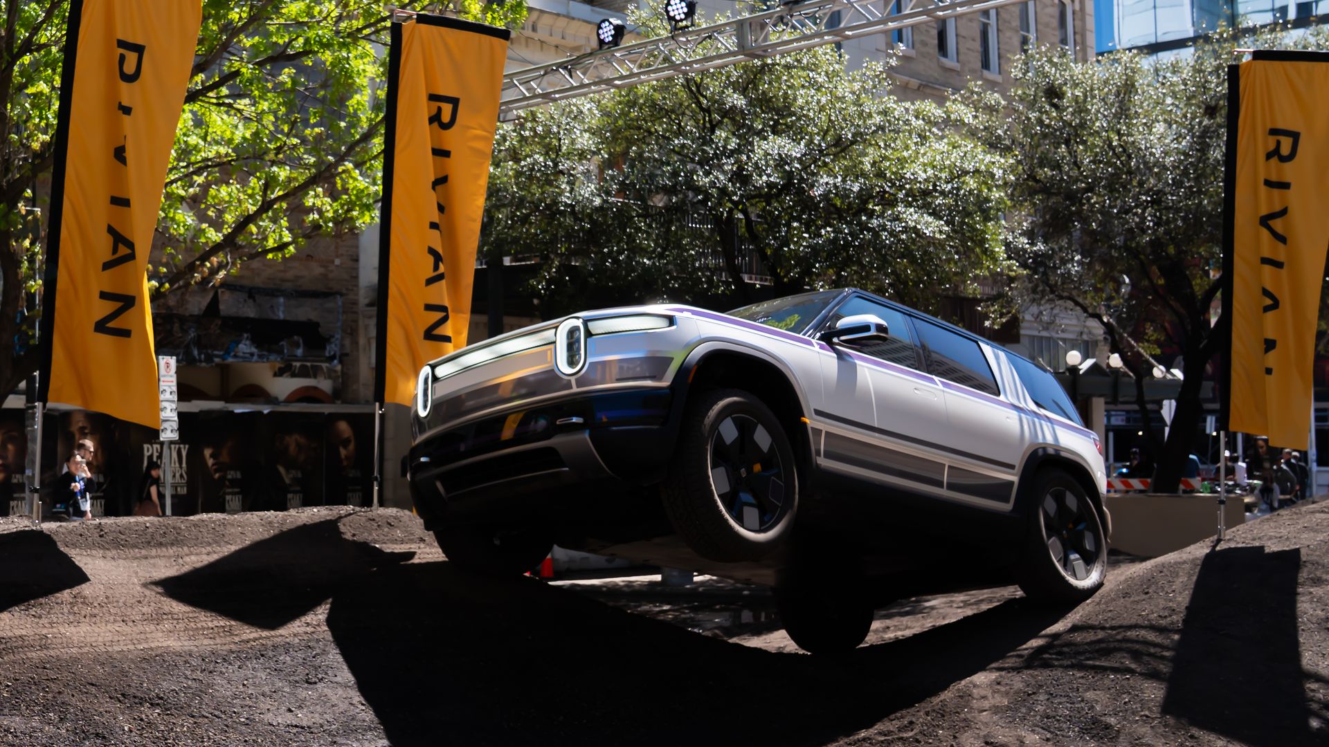 A silver Rivian truck on a raised dirt platform at an outdoor event, flanked by tall yellow banners reading "RIVIAN", with a metal truss and stage lights overhead and green trees in the background.