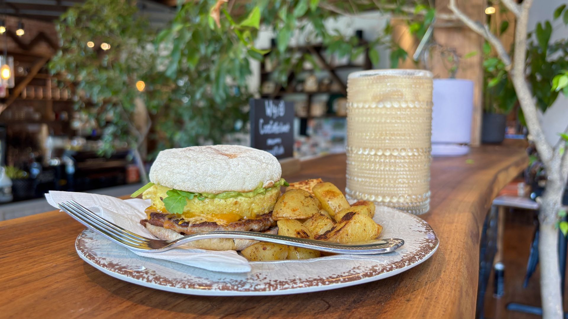 Burger with cheese and lettuce on a sesame bun, with potato wedges on a white plate. A beige drink jar sits on a wooden table; blurred cafe greenery and a chalkboard sign in the background.
