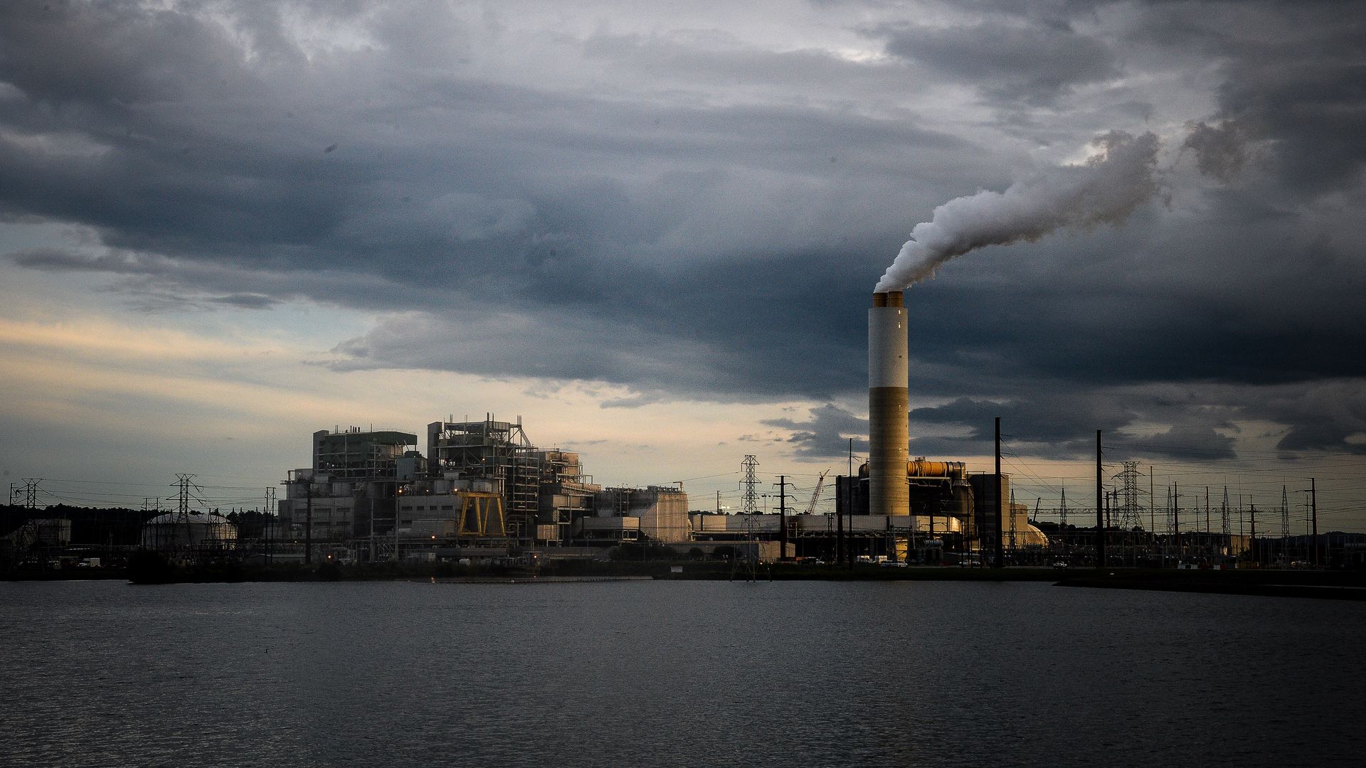 Power plant near water under dark, cloudy sky with smoke billowing.