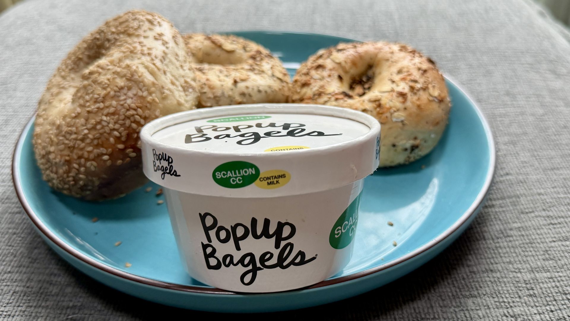 Turquoise plate holds a sesame-seed bagel and an onion-topped bagel, with a Pop-Up Bagels scallion cream cheese tub in the foreground; gray tablecloth and a window in the background.