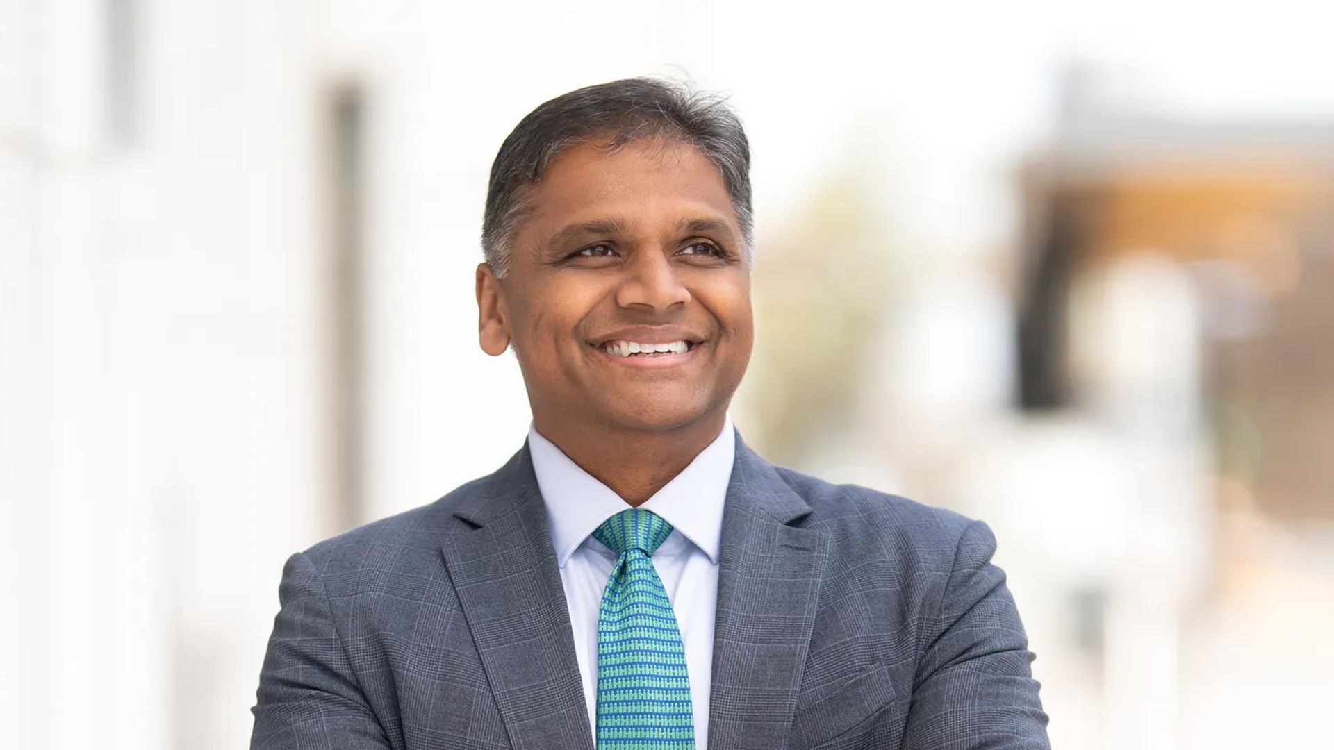 Danny Avula smiling in front of a blurred background in a suit with a teal tie.