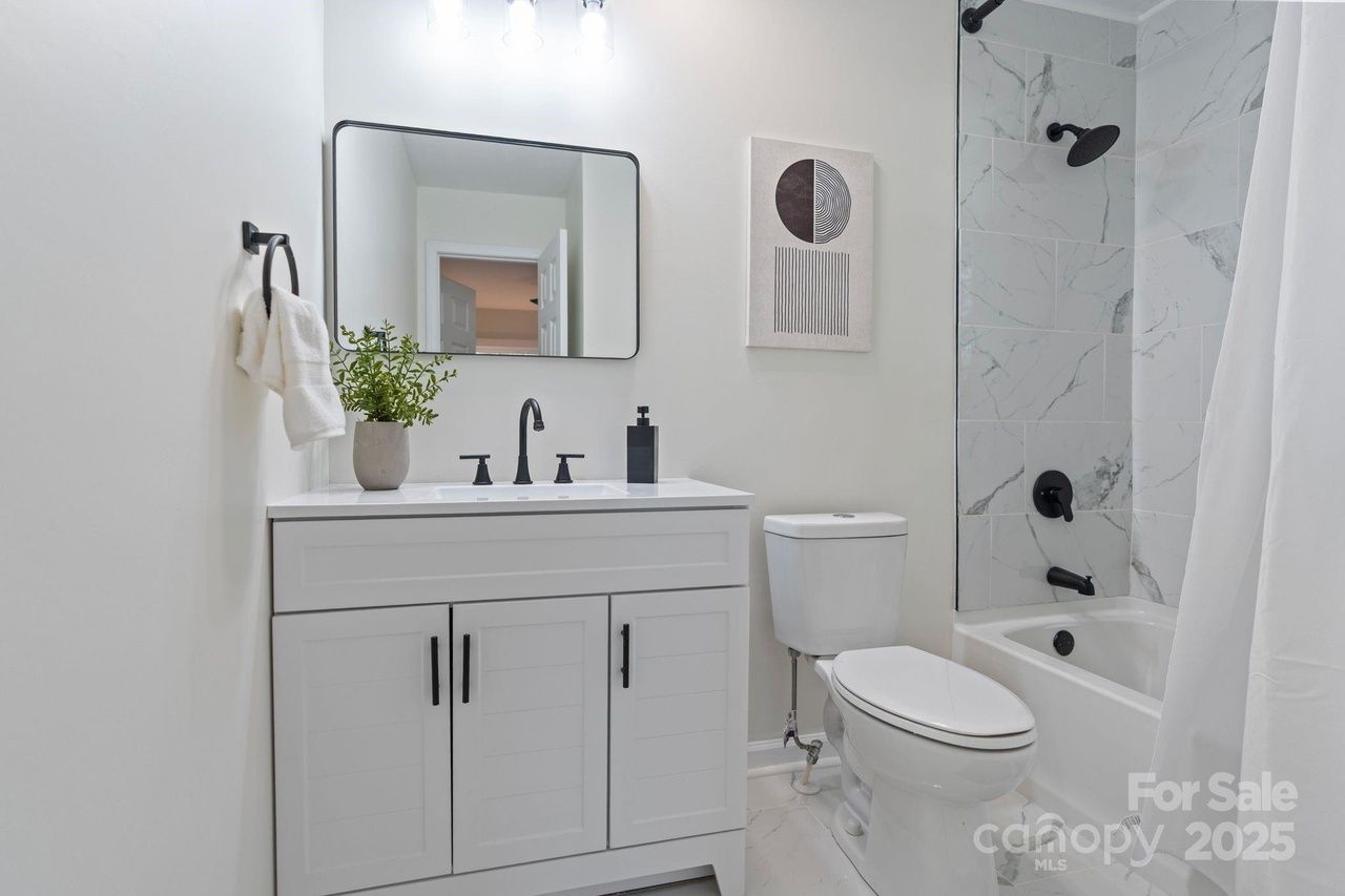 Modern white bathroom with black fixtures, white vanity with plant, rectangular mirror, toilet, and marble-tiled shower with white curtain.
