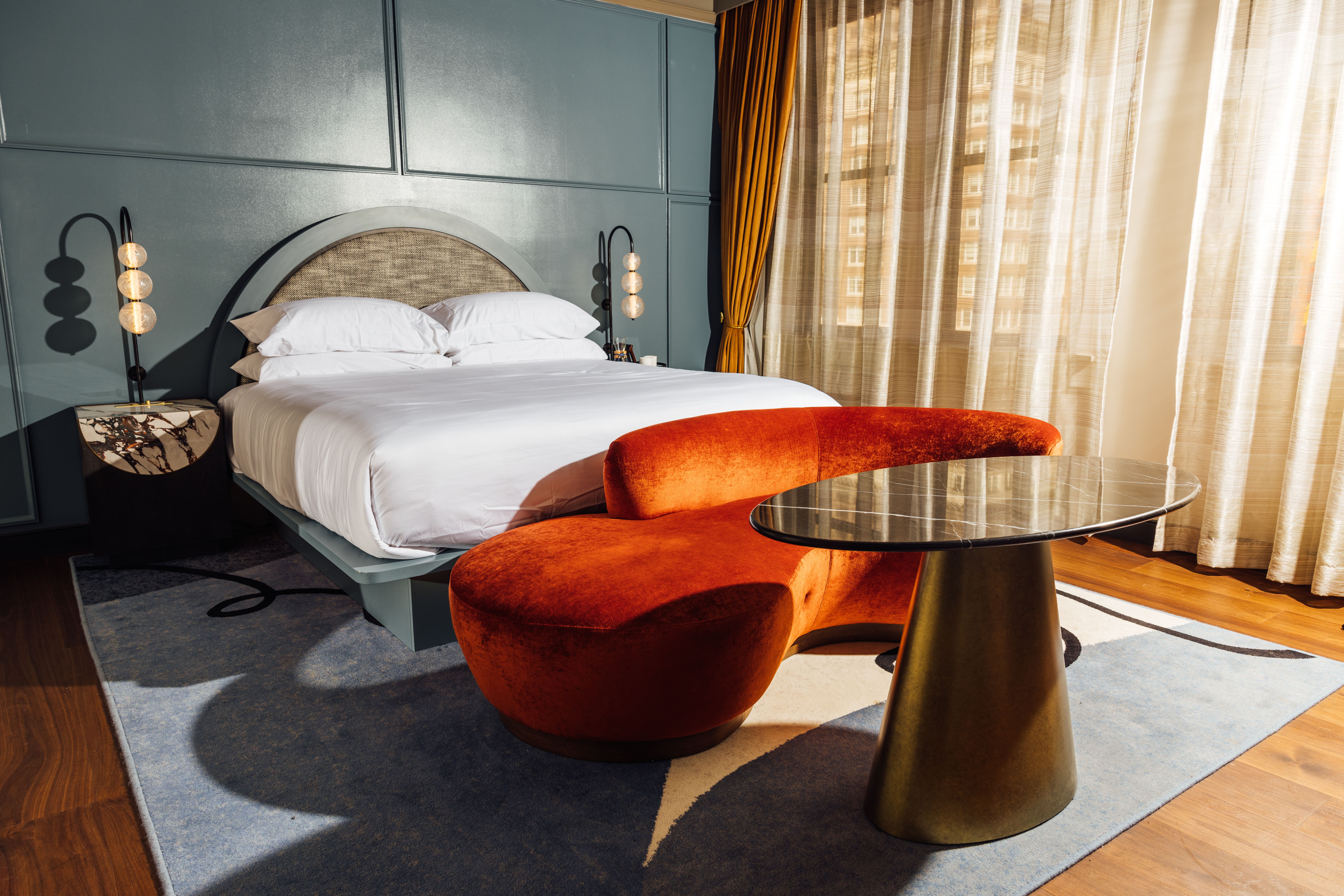 Hotel room with a white bed, blue-gray paneled wall, orange velvet curved sofa, marble-top table, beige curtains, and wooden floor with a blue patterned rug.