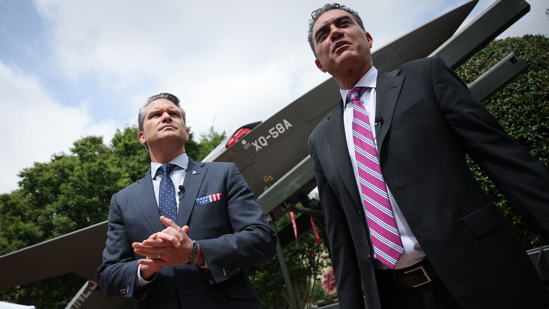 Two men in suits stand outside in front of a large drone and rail launcher. They are in suits and ties. The sun is out.