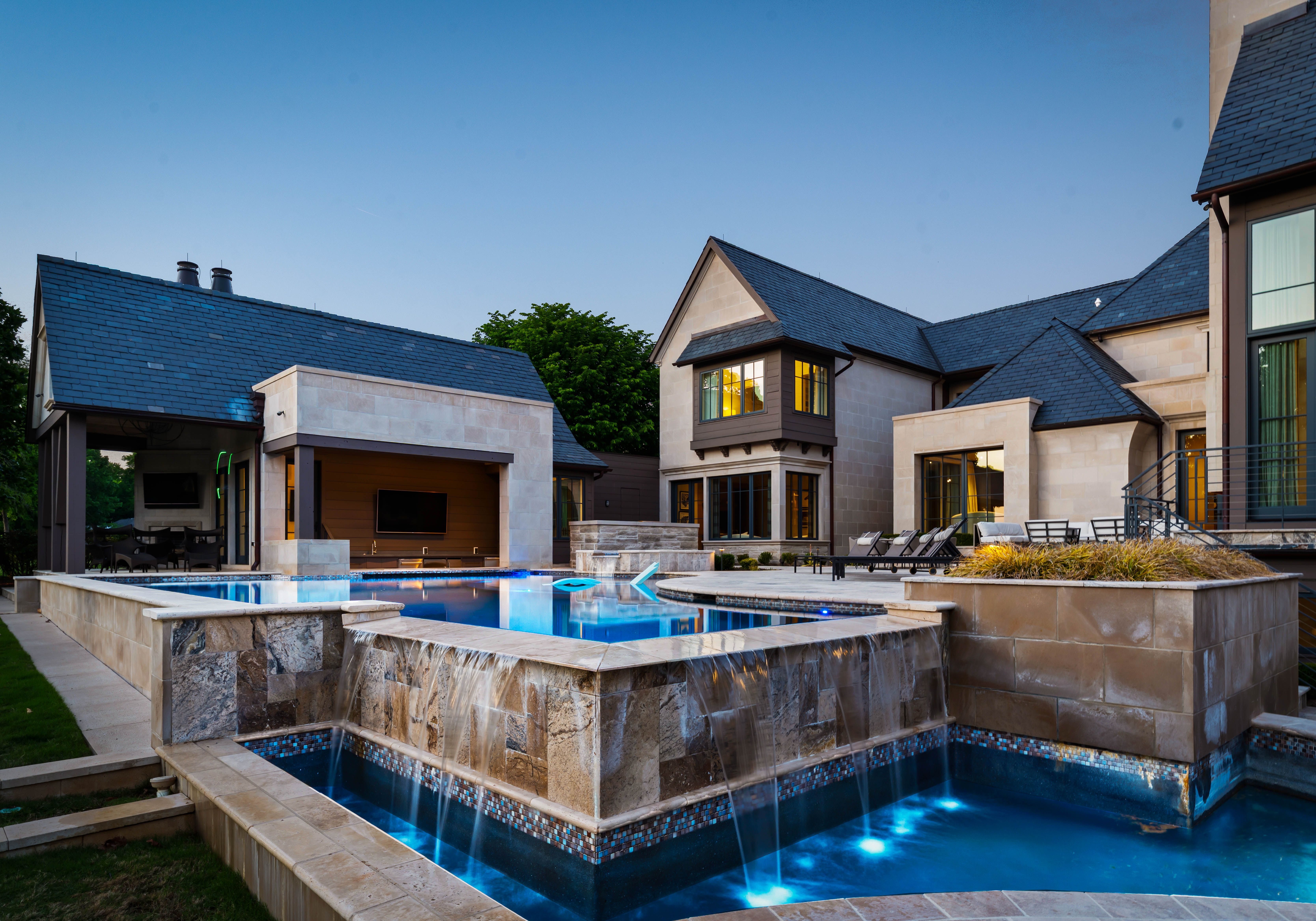Modern mansion with blue slate roofs and beige stone walls surrounding a rectangular pool with blue lighting, outdoor seating, and lush trees under a clear blue sky.