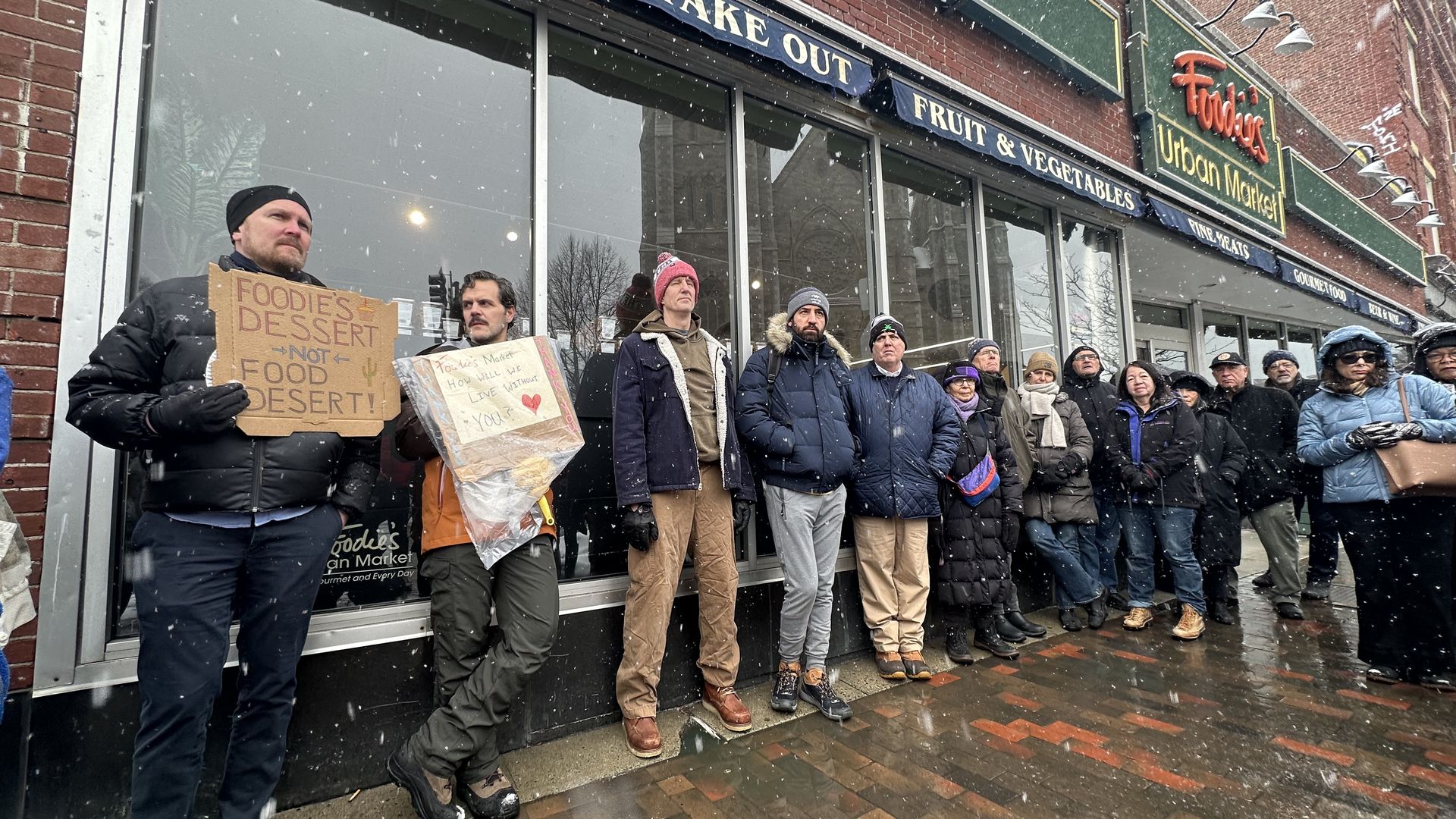 South End residents stand outside Foodie's Market in the snow Saturday morning, protesting plans to convert the grocery store for another use. Two attendees hold signs saying "Foodie's dessert, not food desert!" and "Foodie's Market, how will we live without you?"