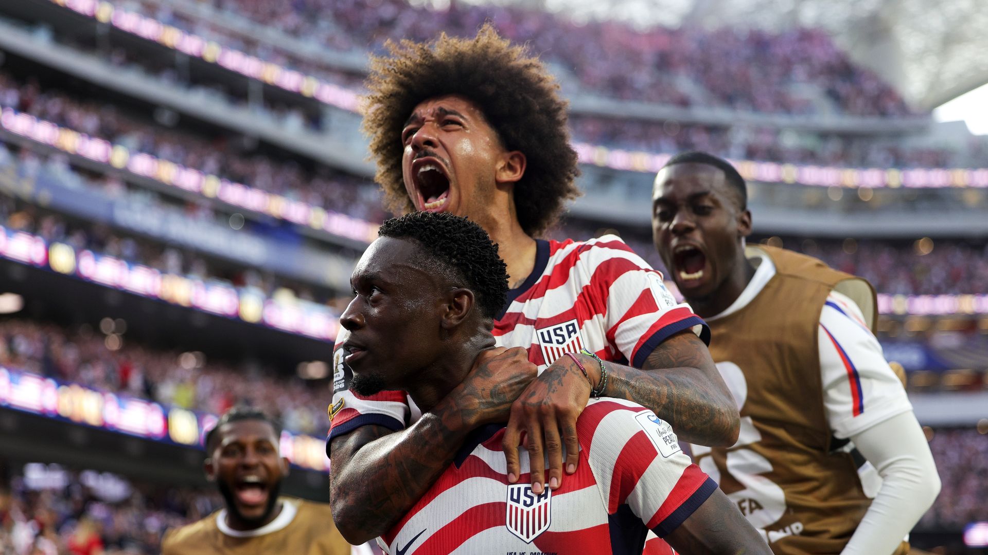 USA soccer players celebrate on the field; center player with an Afro screams in excitement while teammates hug him, stadium crowd blurred in the background.