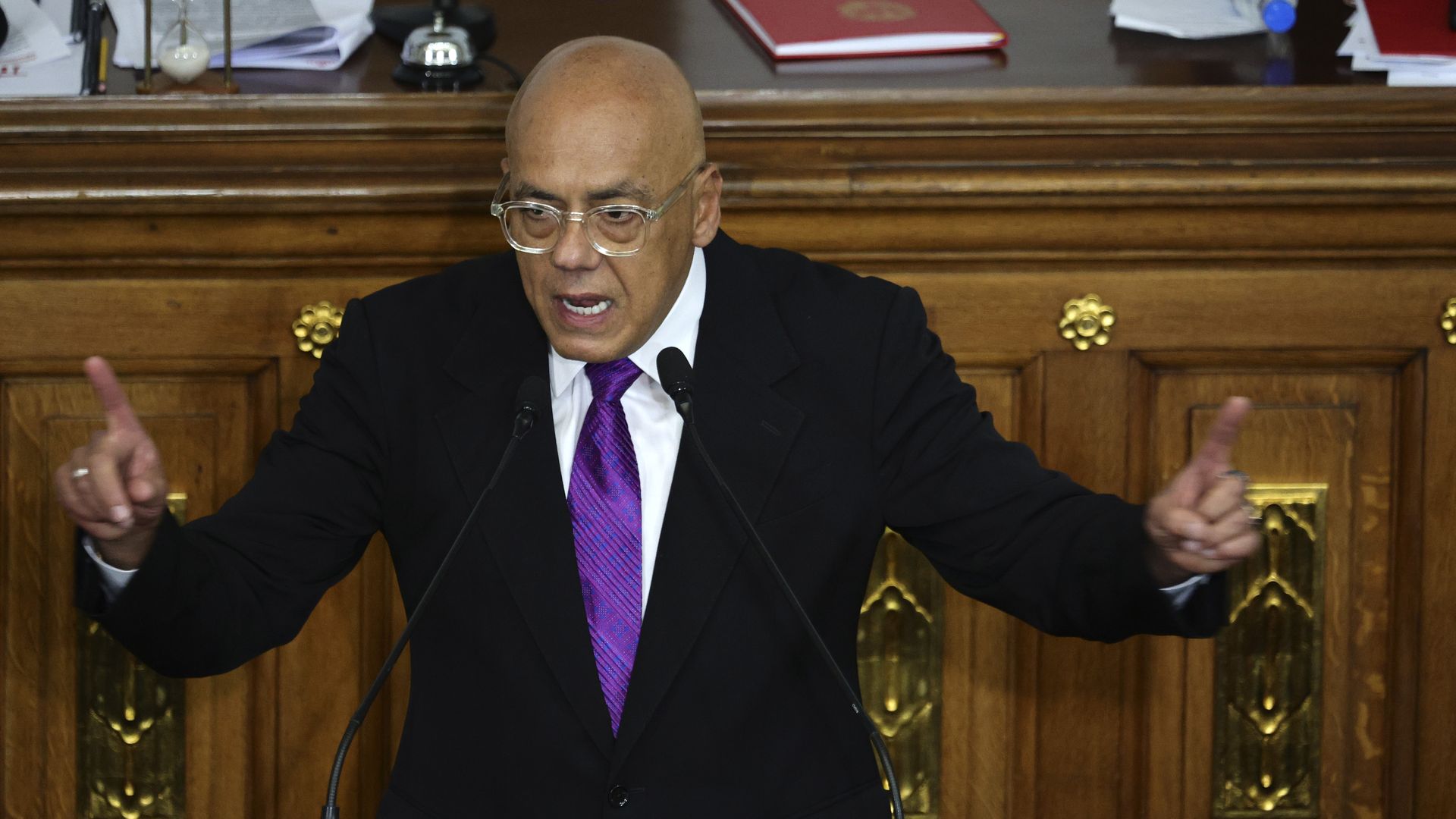 Rodríguez speaking into two microphones while holding up both of his index fingers wearing a black suit jacket, white collared shirt and purple tie 