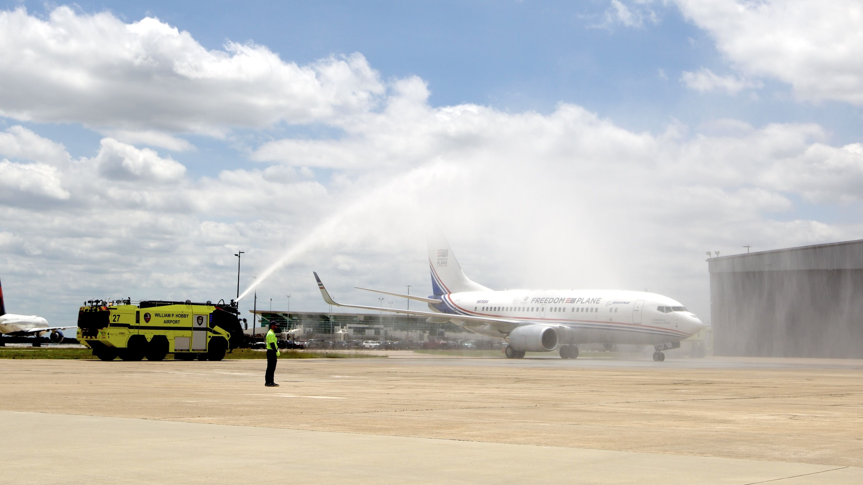 Yellow fire truck sprays a water arch over a white passenger plane on the tarmac; a worker in neon vest stands nearby at William P. Hobby Airport, blue sky and clouds overhead.