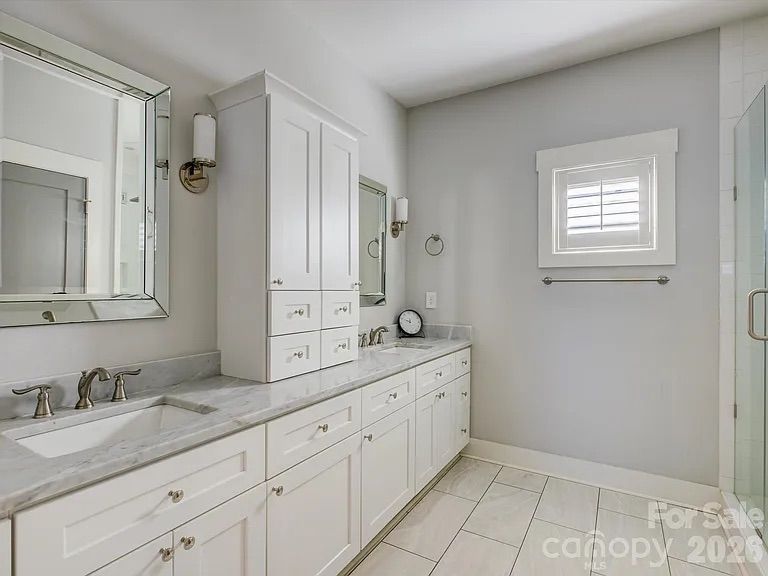 Modern bathroom with white cabinetry, marble countertop, two mirrors, silver fixtures, tiled floor, small window with white shutters, and a glass shower door.