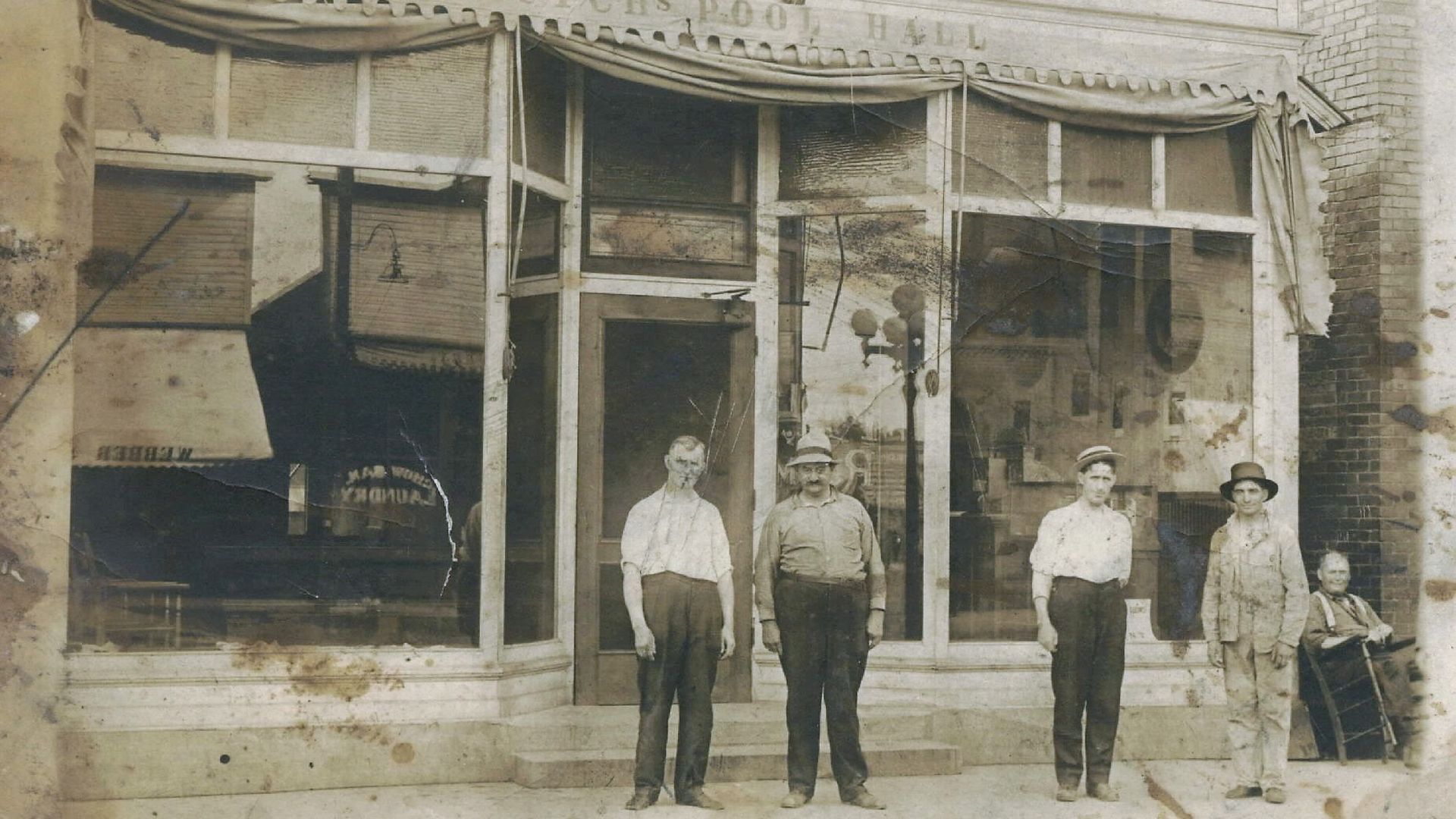 Sepia photo of a vintage storefront with large glass windows and a draped awning. A faded sign reads "POOL HALL". Five people in early 20th-century attire stand on the sidewalk.