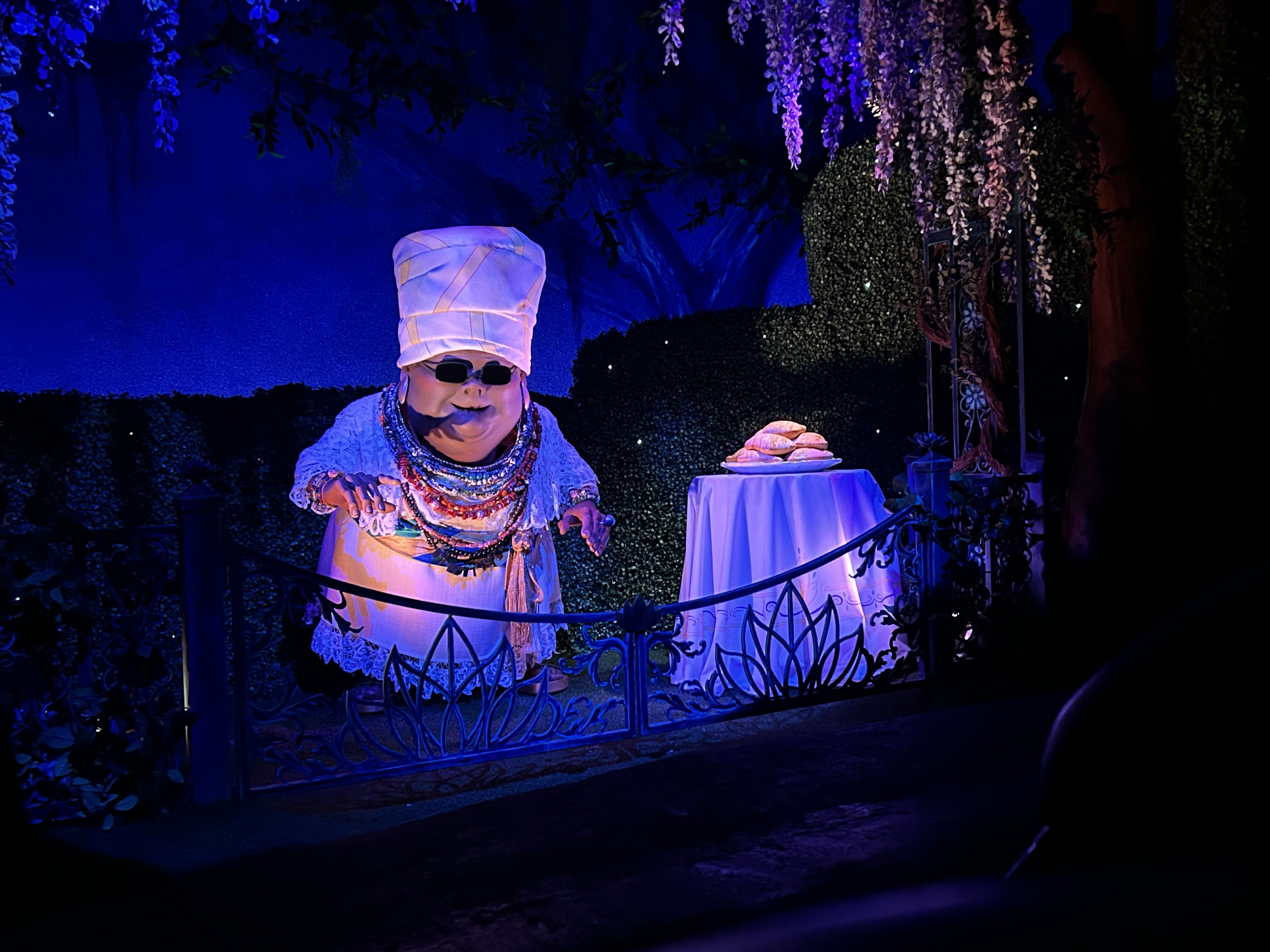 An animatronic version of the Mama Odie character standing next to a table with a plate of beignets. 
