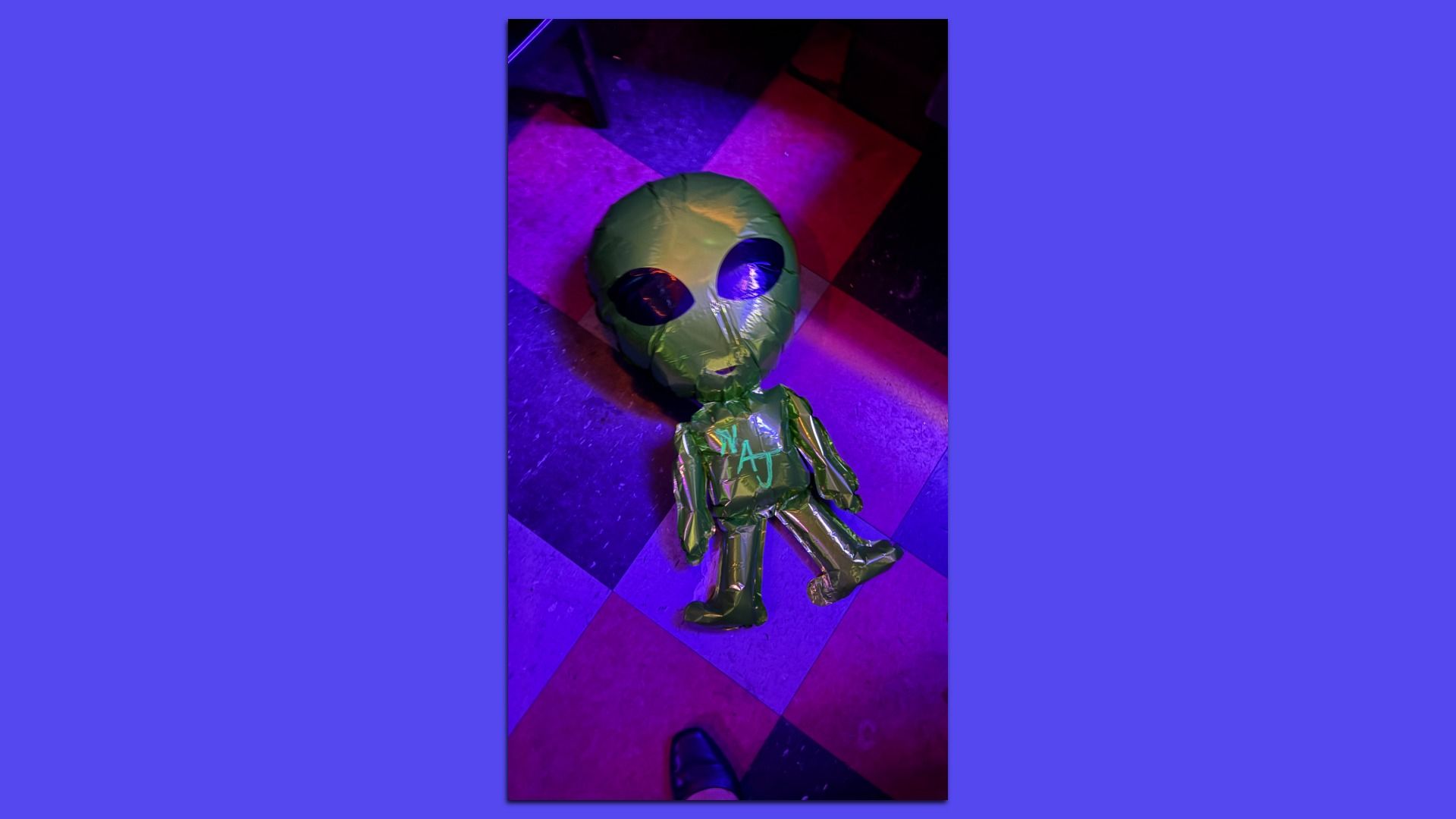 Top-down view of a green inflatable alien toy with large black eyes, lying on a purple and pink checkered floor under neon purple light; a shoe is visible at the bottom edge.