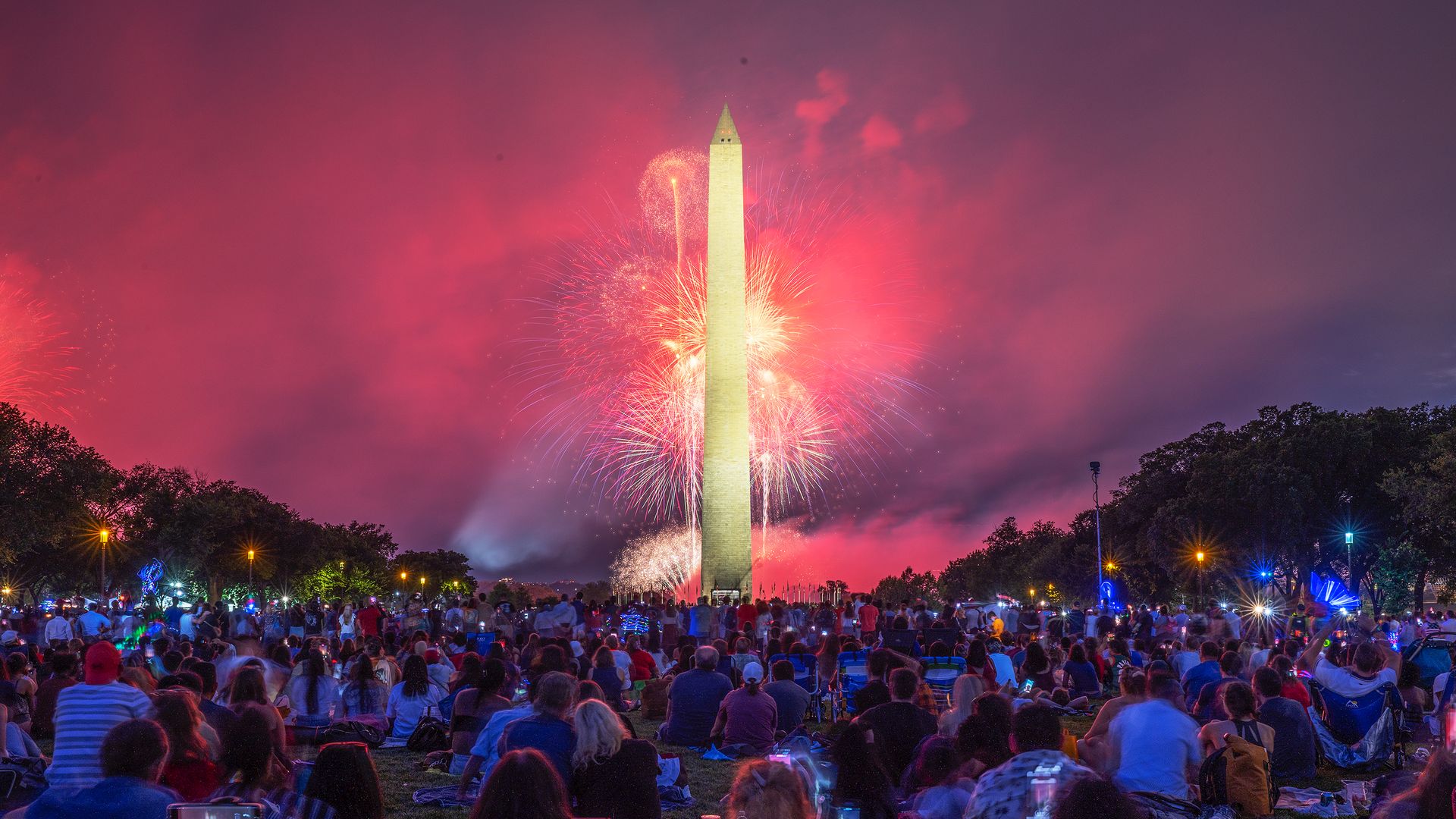 The Washington monument is in the background with people sitting in front of it. Fireworks are going off behind the monument.