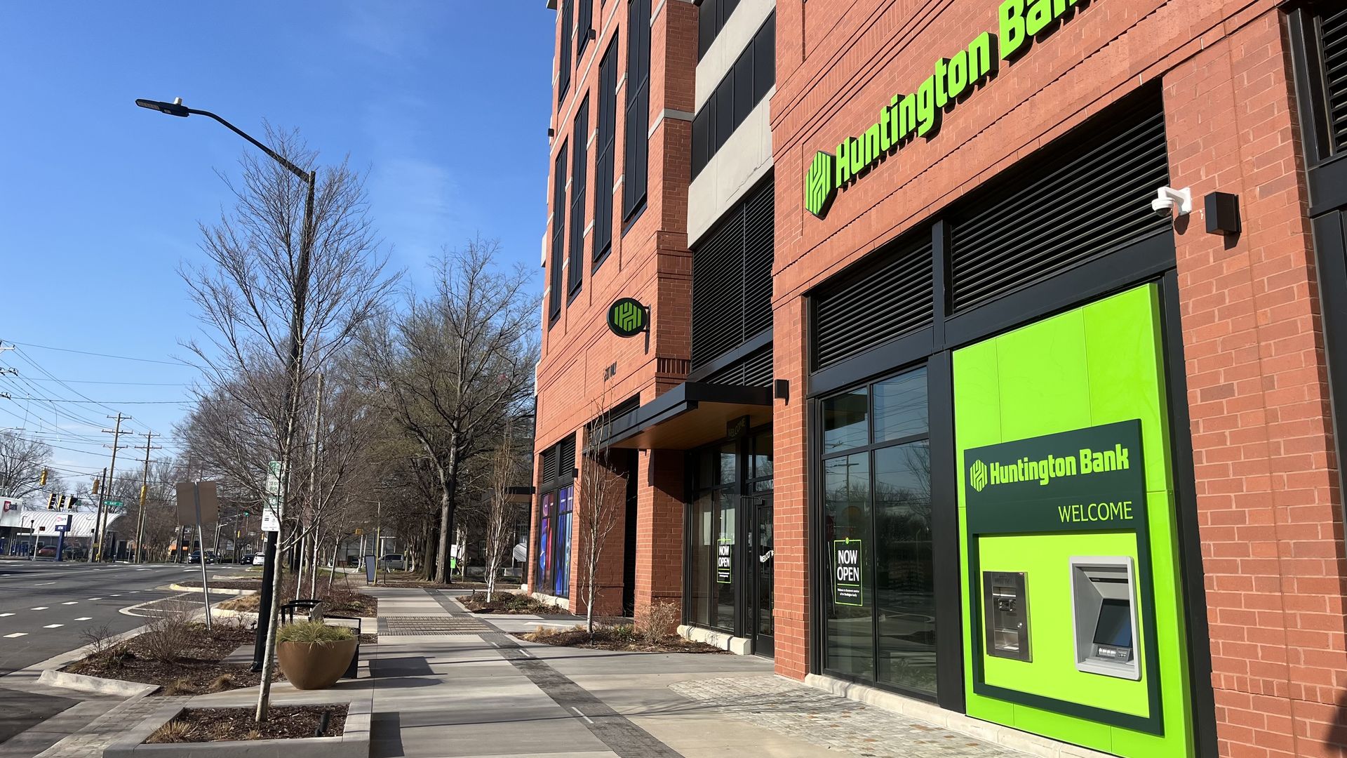 Street view of a red brick Huntington Bank building with bright green signage and ATM, clear blue sky, bare trees, and a clean sidewalk in daytime.