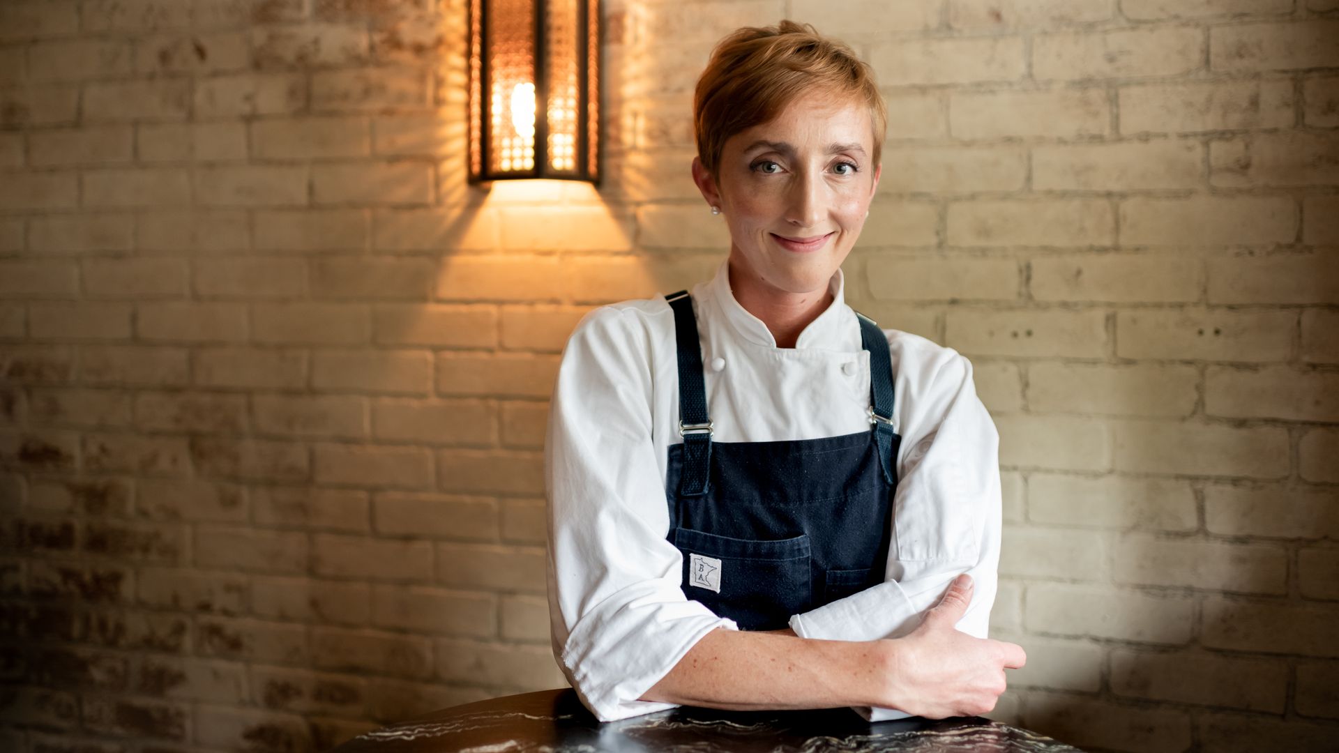 A woman with light skin and reddish hair in a white chef's uniform with a black apron poses for a photograph