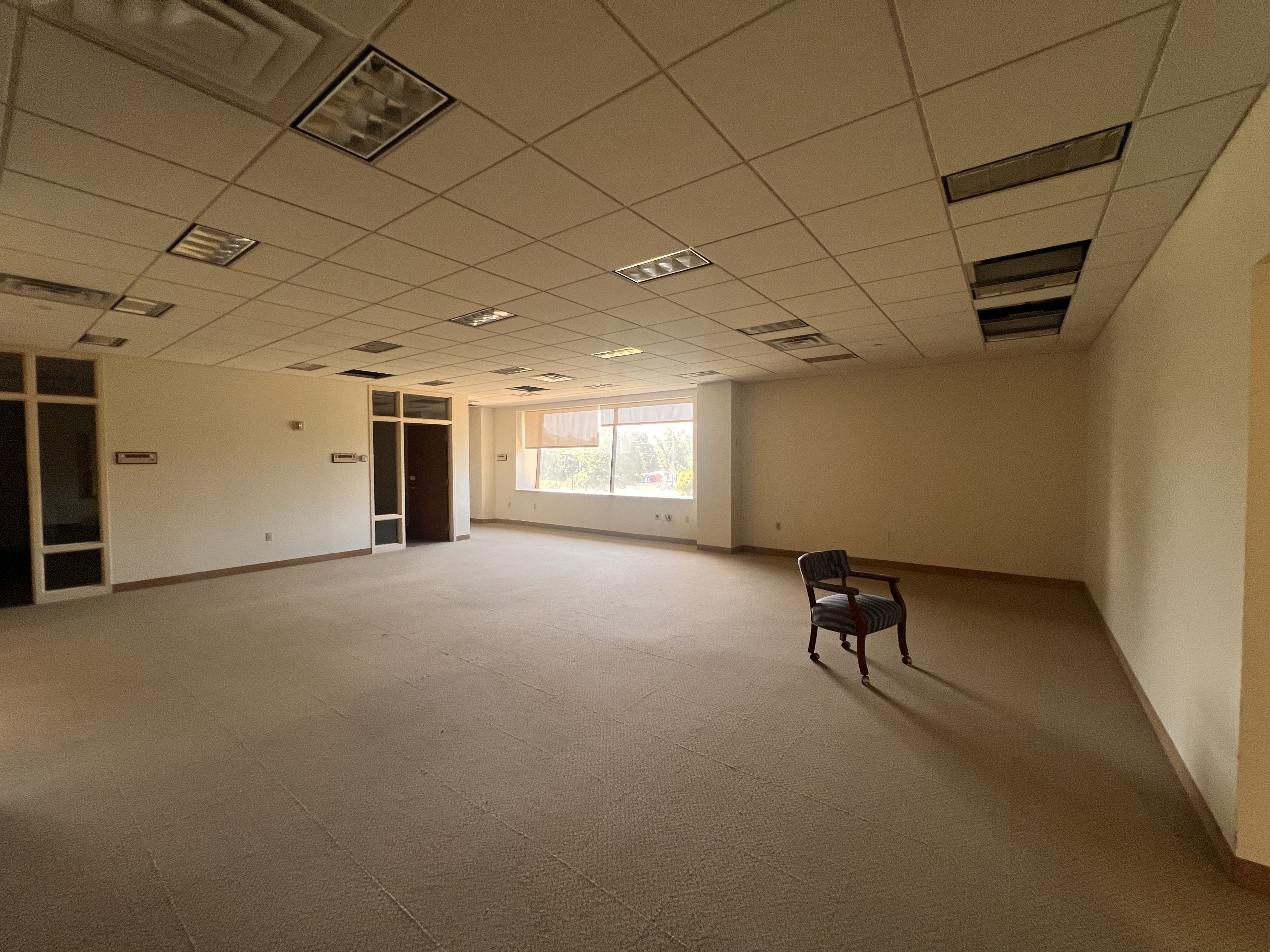 An empty office room with a tiled ceiling and fluorescent lights. Beige walls, carpeted floor, a few chairs in the foreground, and large windows at the far end.