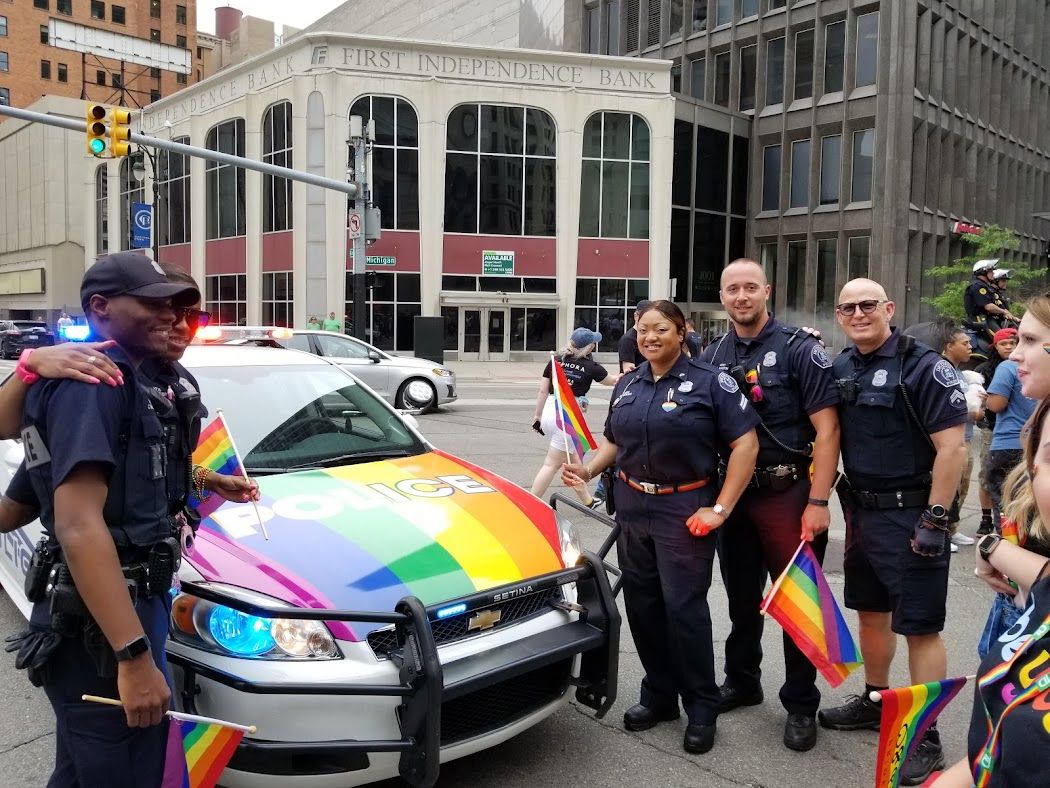 Police officers with flags stand next to a police car with a rainbow wrap.