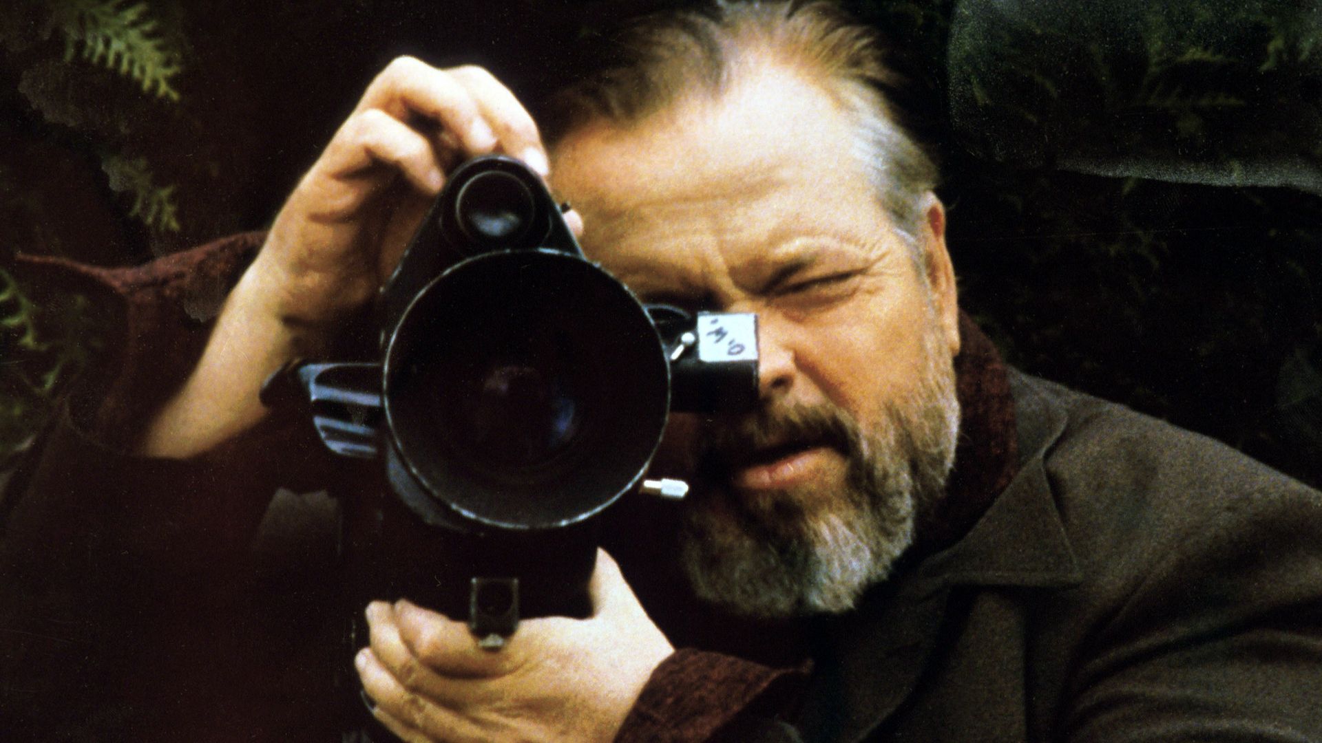 A bearded man in a brown coat uses a large vintage movie camera, looking through the viewfinder outdoors amid green foliage.