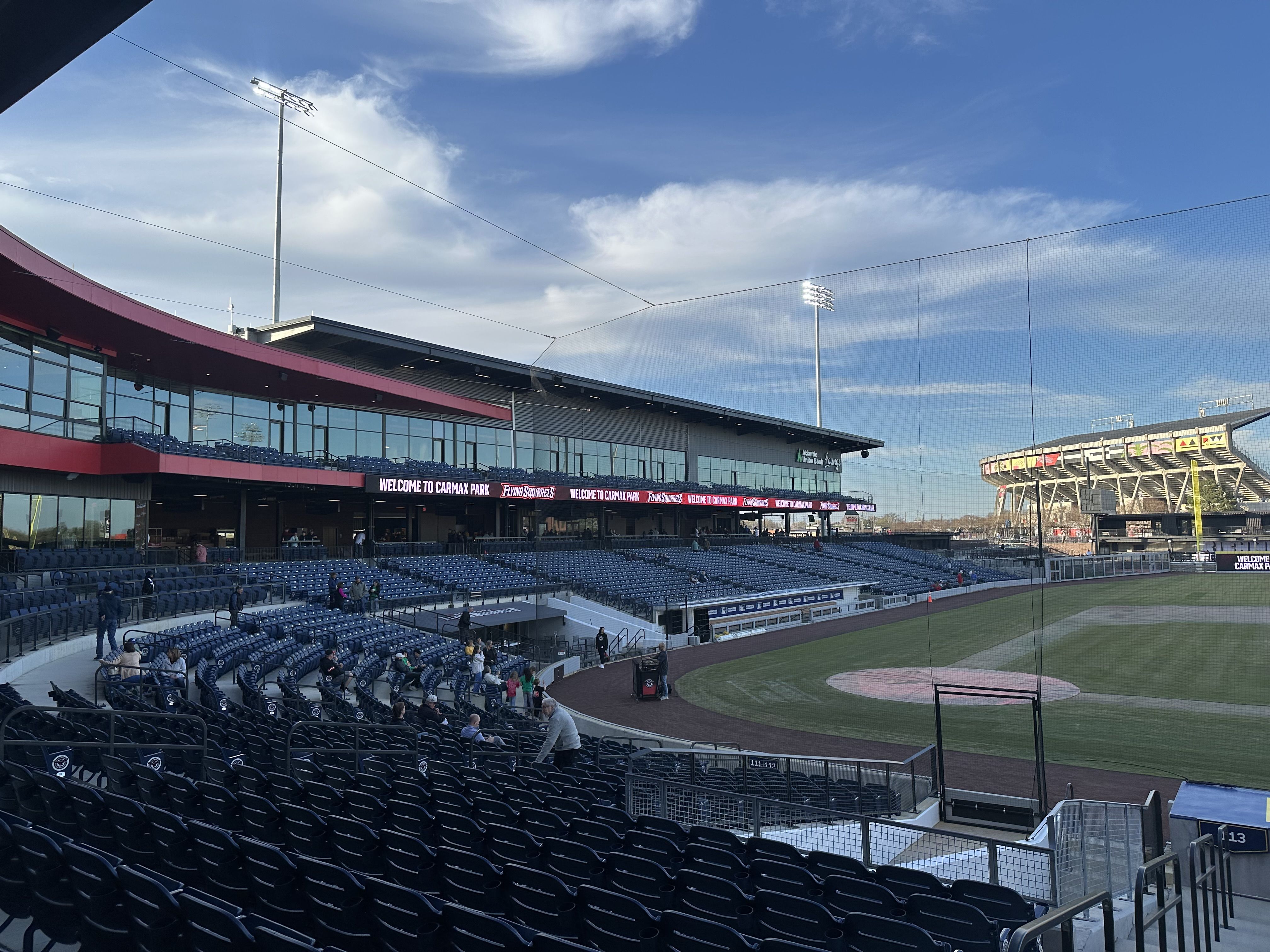 Wide view of a sunny baseball stadium with blue seats, a red-trimmed concourse, banners reading "Welcome to CarMax Park," a dirt infield, and tall light towers against a clear blue sky.