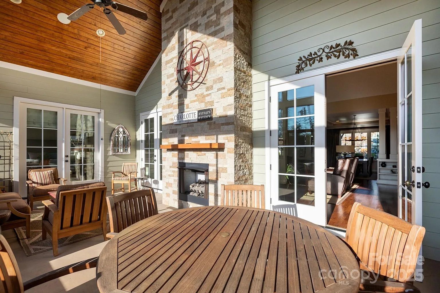 Sunlit patio with wooden ceiling, ceiling fan, round wooden table and chairs, cushioned seating, stone fireplace with compass rose decor, and glass doors leading indoors.