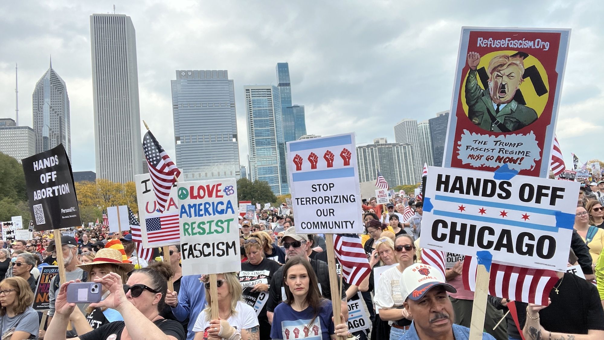 Crowd of protesters in Chicago with signs including "HANDS OFF CHICAGO," "STOP TERRORIZING OUR COMMUNITY," "LOVE AMERICA RESIST FASCISM," and a caricature of Trump with "The Trump Fascist Regime Must Go Now!" in front of Chicago skyline.