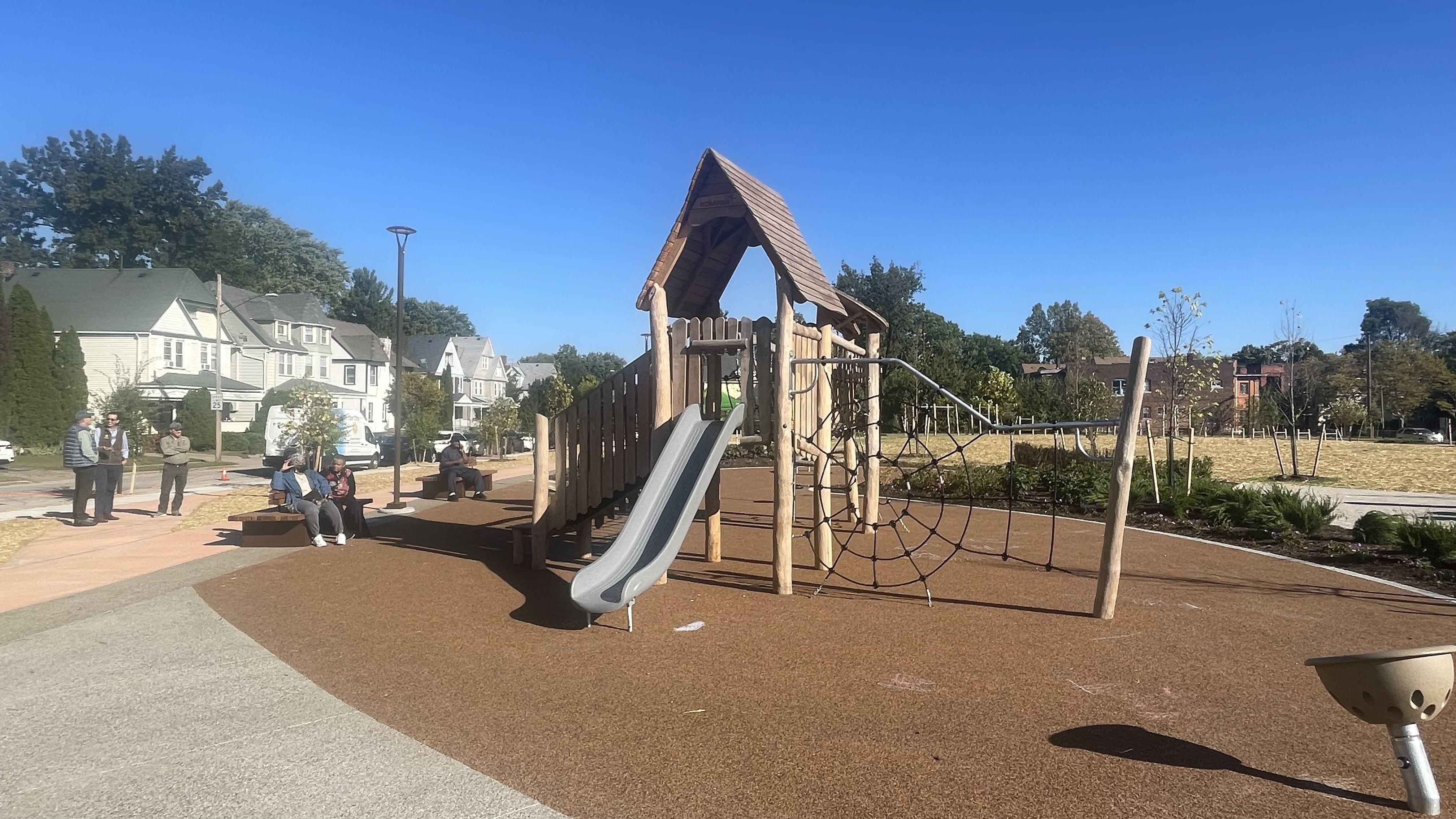 Wooden playground structure with slide and rope climbing net on brown soft flooring in a sunny park, with people sitting on benches and houses in the background under a clear blue sky.