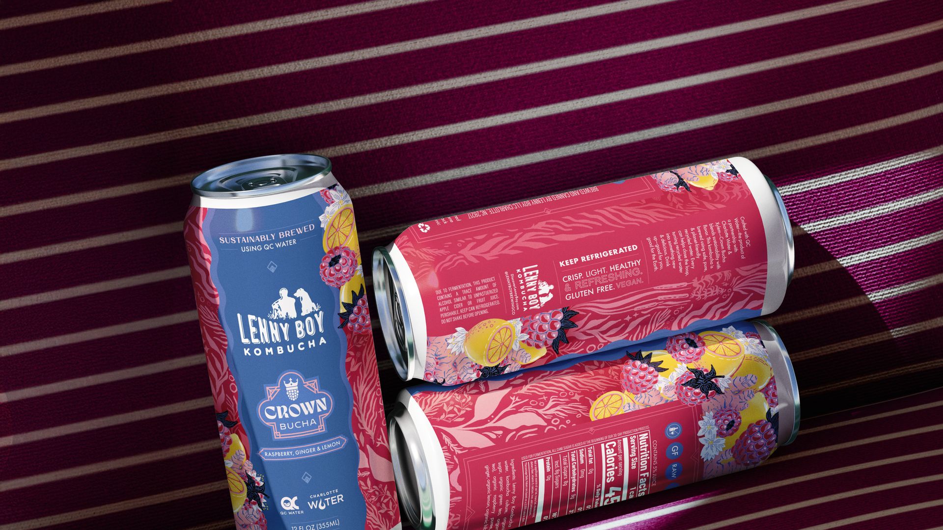 Three cans of Lenny Boy Kombucha Crown Bucha with raspberry, ginger & lemon flavor on a maroon and white striped surface with floral and fruit illustrations on cans.