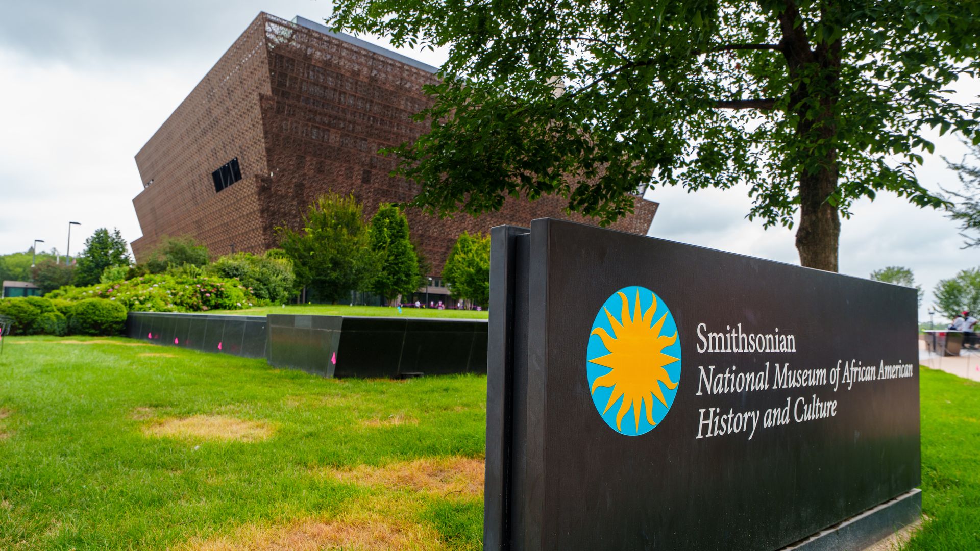 Black sign with the Smithsonian logo and text for the National Museum of African American History and Culture, with the museum building and green trees in the background.