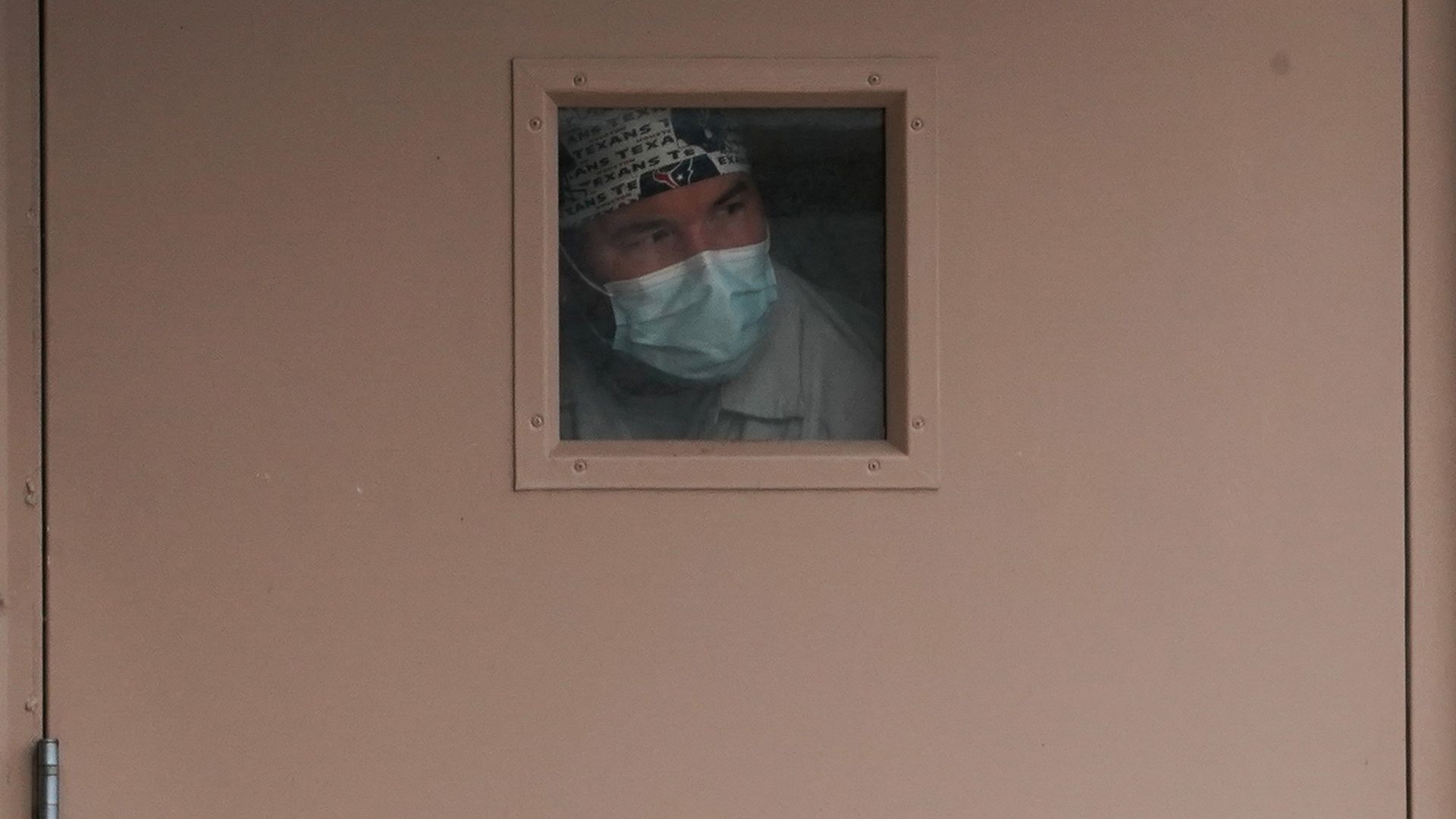 In this image, a man wearing a face mask stands in front of a tiny window.