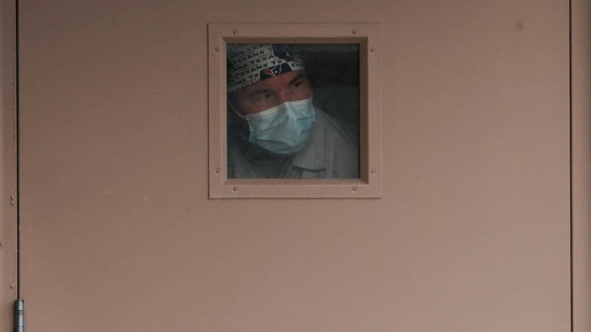 In this image, a man wearing a face mask stands in front of a tiny window.