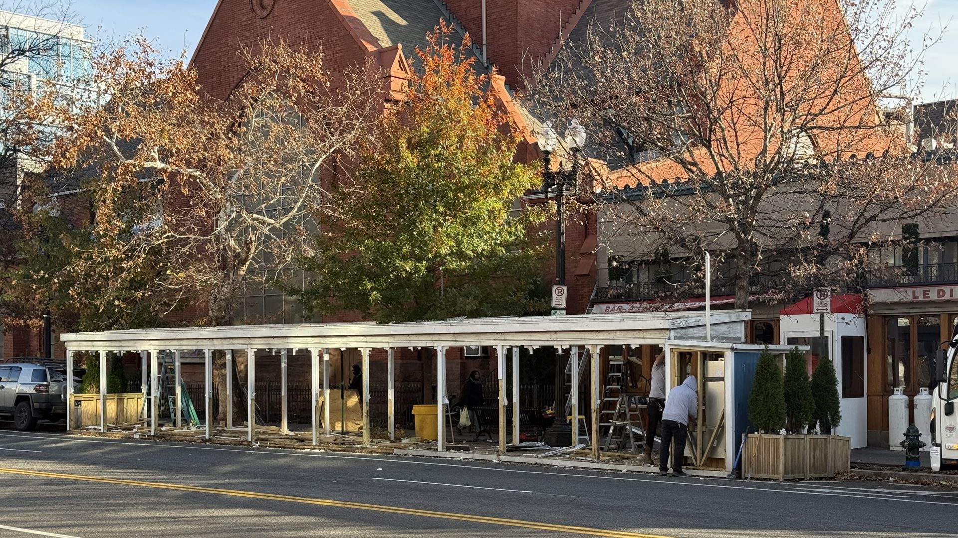 Long white outdoor structure under construction on a city street, with workers and ladders, autumn trees with orange and green leaves, and a red brick building in the background.