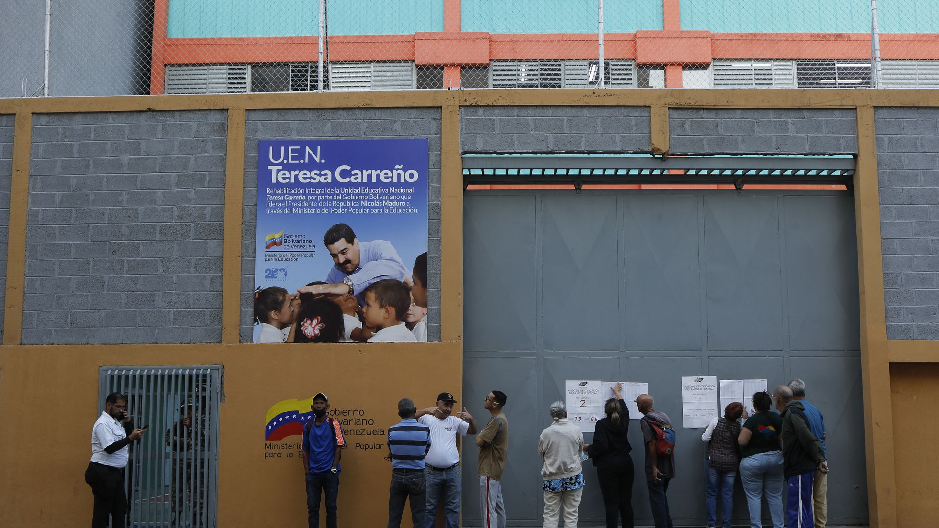 a row of men stand outside a polling station in Venezuela on Sunday. The wall is mustard orange. 