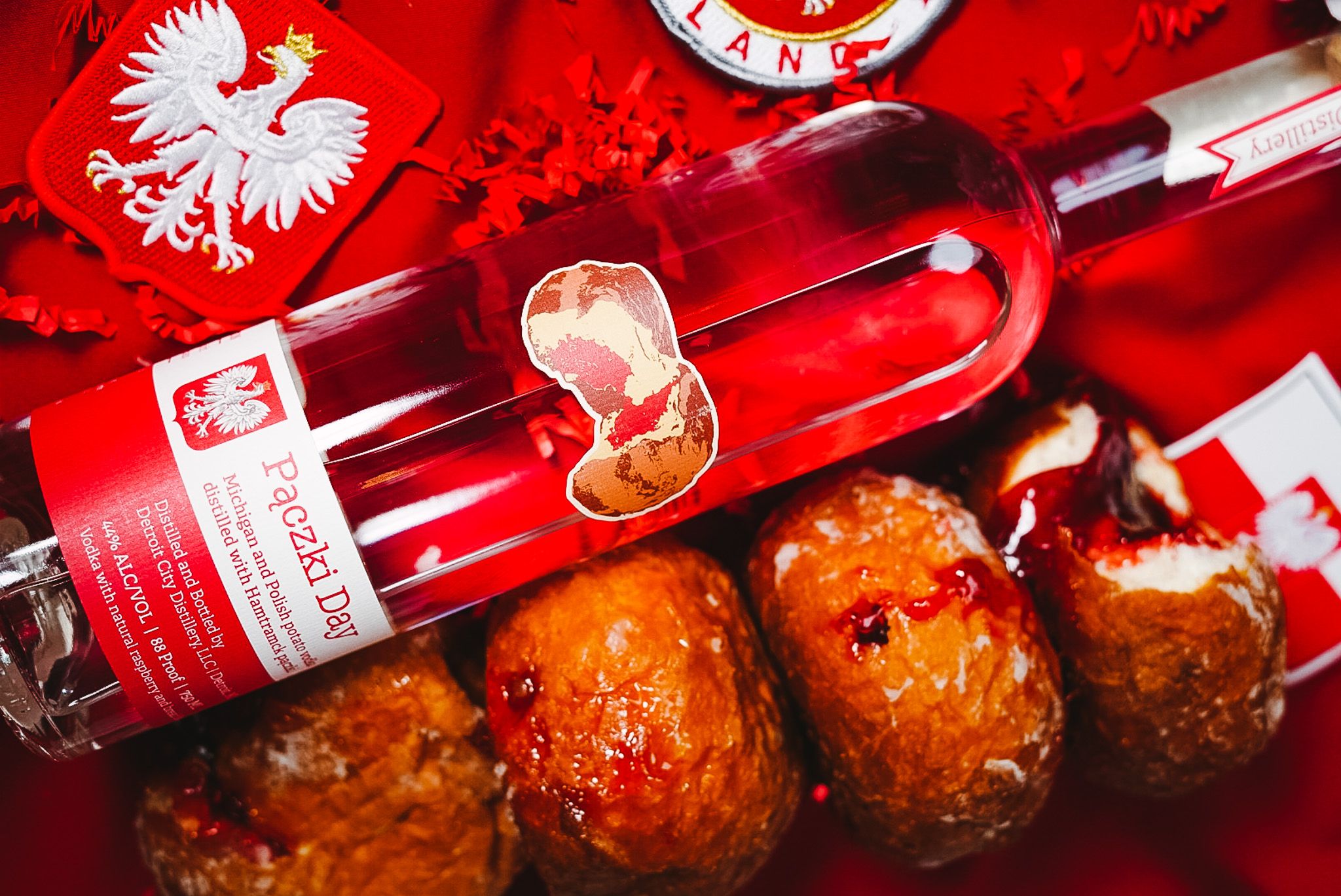 Bottle of Paczki Day vodka with a red label featuring a white eagle, surrounded by four jelly-filled paczki pastries on a red background with red decorations.