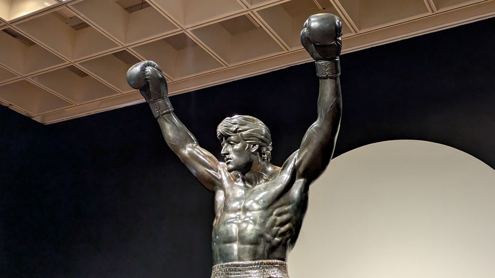 Bronze statue of a muscular boxer with arms raised in victory, wearing boxing gloves and trunks, displayed indoors against a dark wall and a pale circular backdrop.