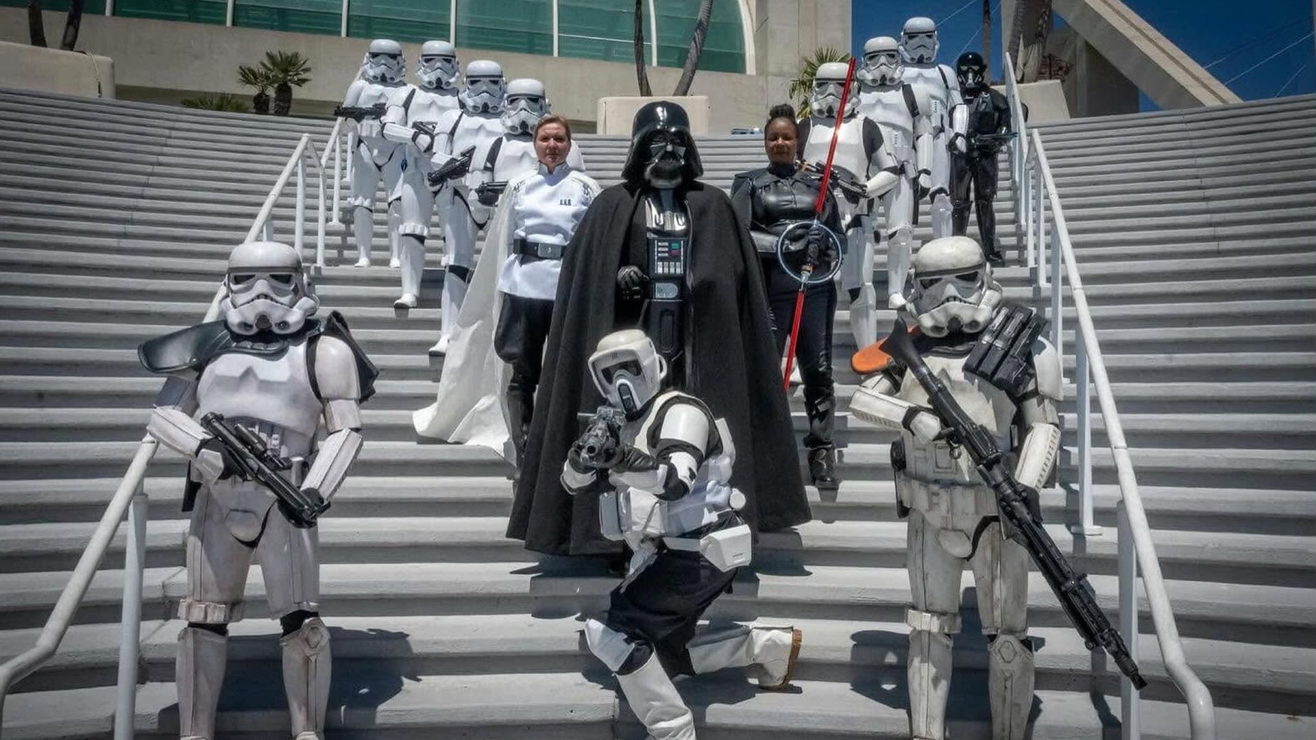 Group of Star Wars cosplayers on outdoor stairs: Darth Vader center in cape, surrounded by white Stormtroopers and two allied characters; some hold blasters, a red lightsaber visible.