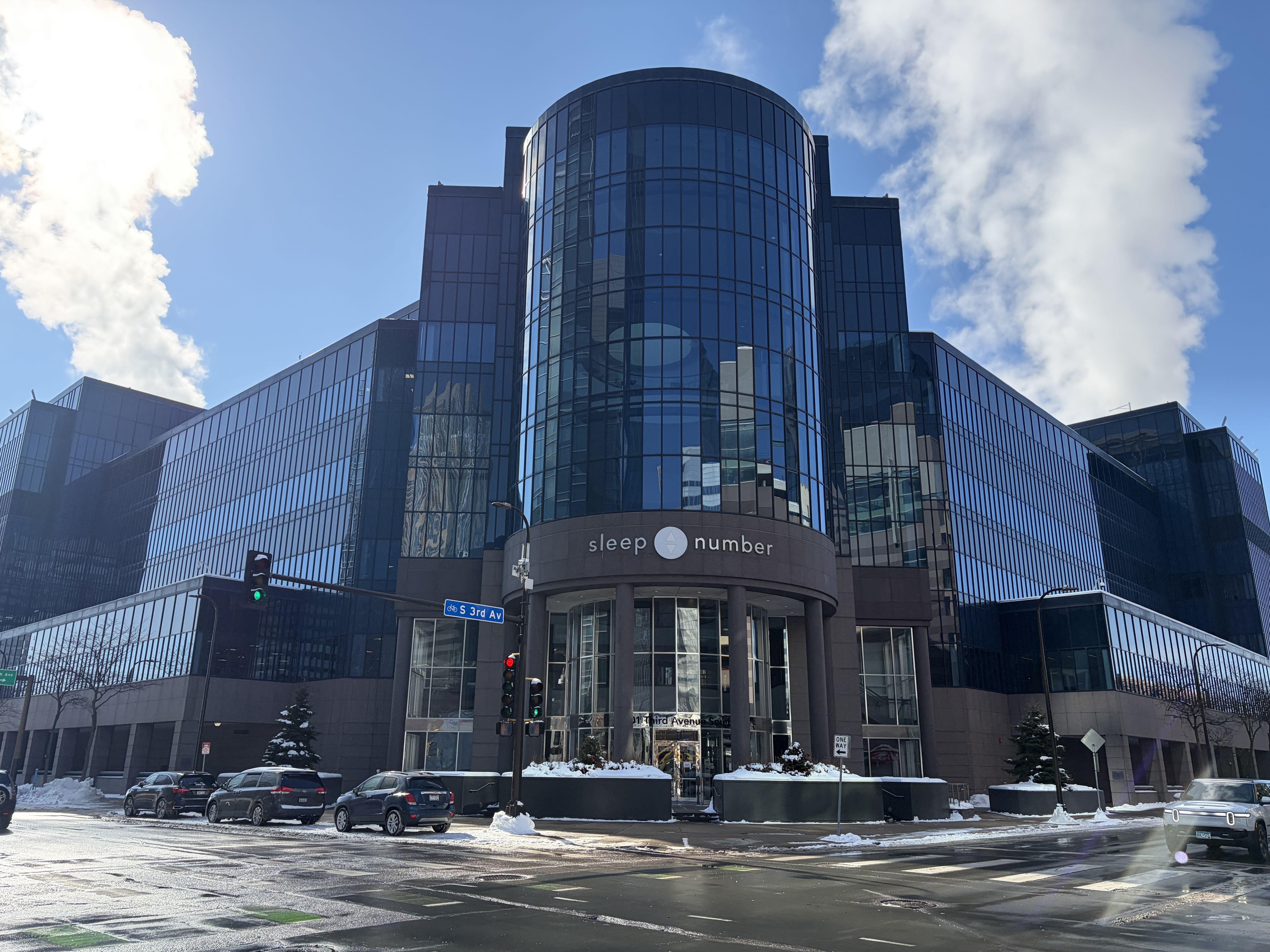 Glass-front office building with a rounded central tower and a Sleep Number sign at the entrance. Snowy street, parked cars, and a blue sky with white clouds above the modern glass facade.