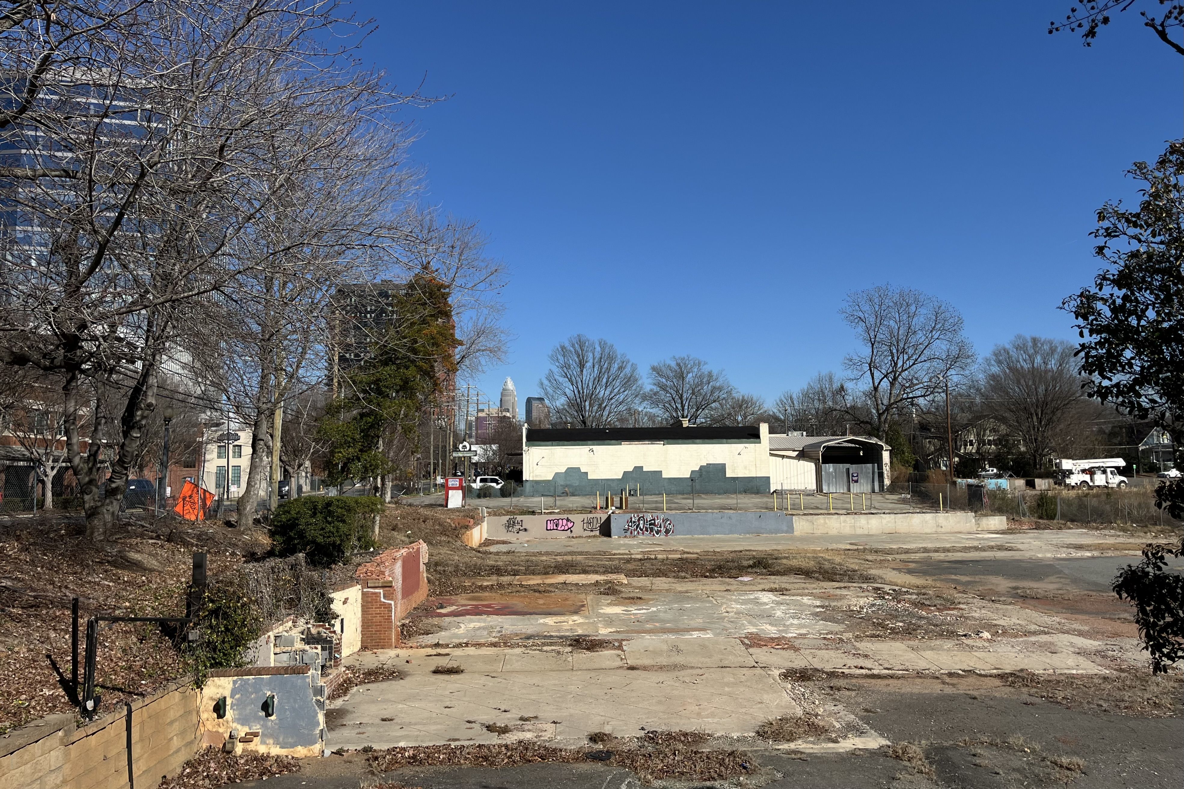 Empty urban lot with concrete foundation remnants, bare trees, graffiti on a low wall, a pale green building, and a clear blue sky in the background.