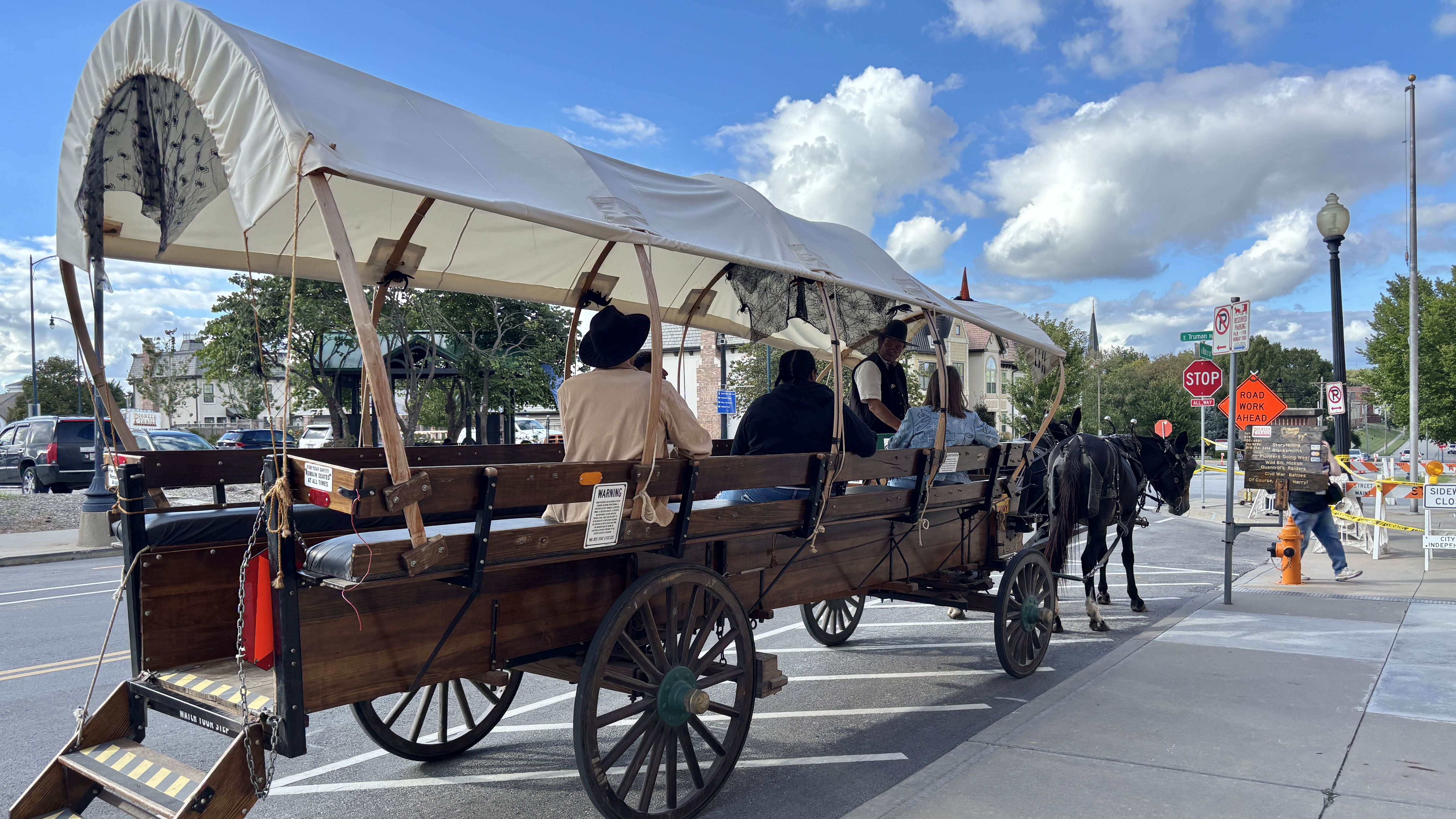 White-covered, wooden horse-drawn wagon with passengers travels along a city street; two black horses pull ahead while a driver in a vest and hat guides them under a bright blue sky.