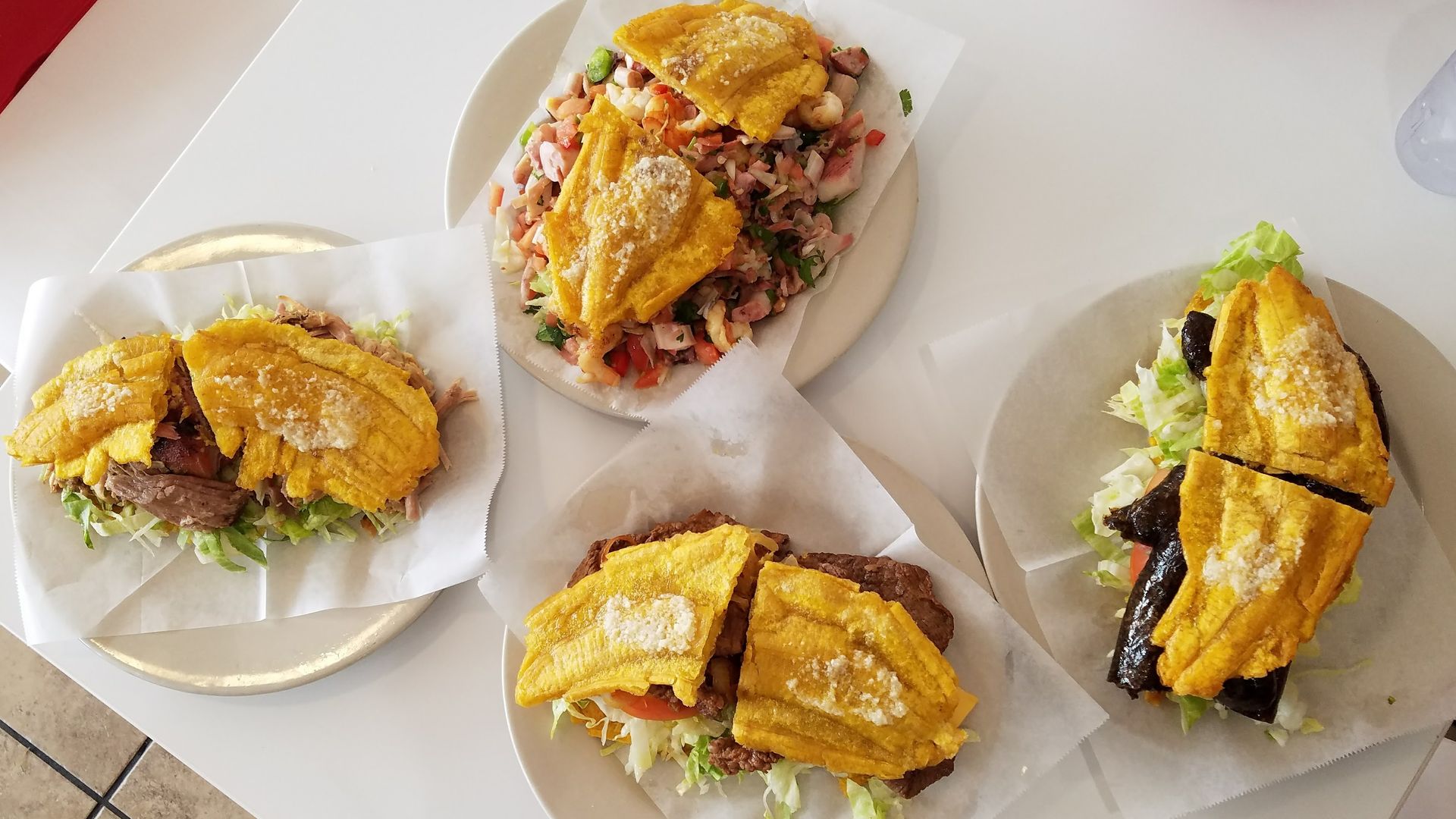 Assorted tacos on white plates with shredded lettuce and diced tomatoes; crispy yellow corn tortilla shells hold meat fillings. A central oval plate shows a chopped salad mix.